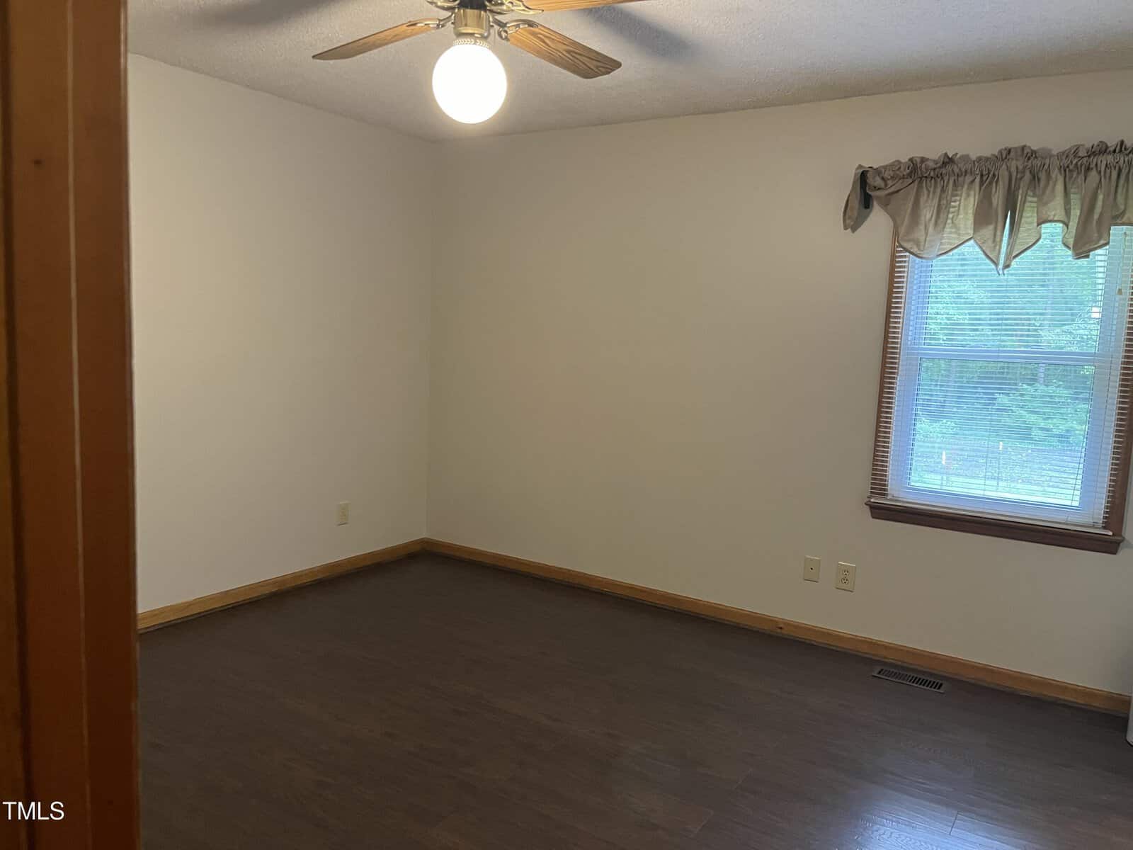 An empty room with dark wooden floors, white walls, and a ceiling fan with a light. A window on the right has a sheer curtain, and natural light is coming through. There's a wooden trim on the floor and around the door frame.