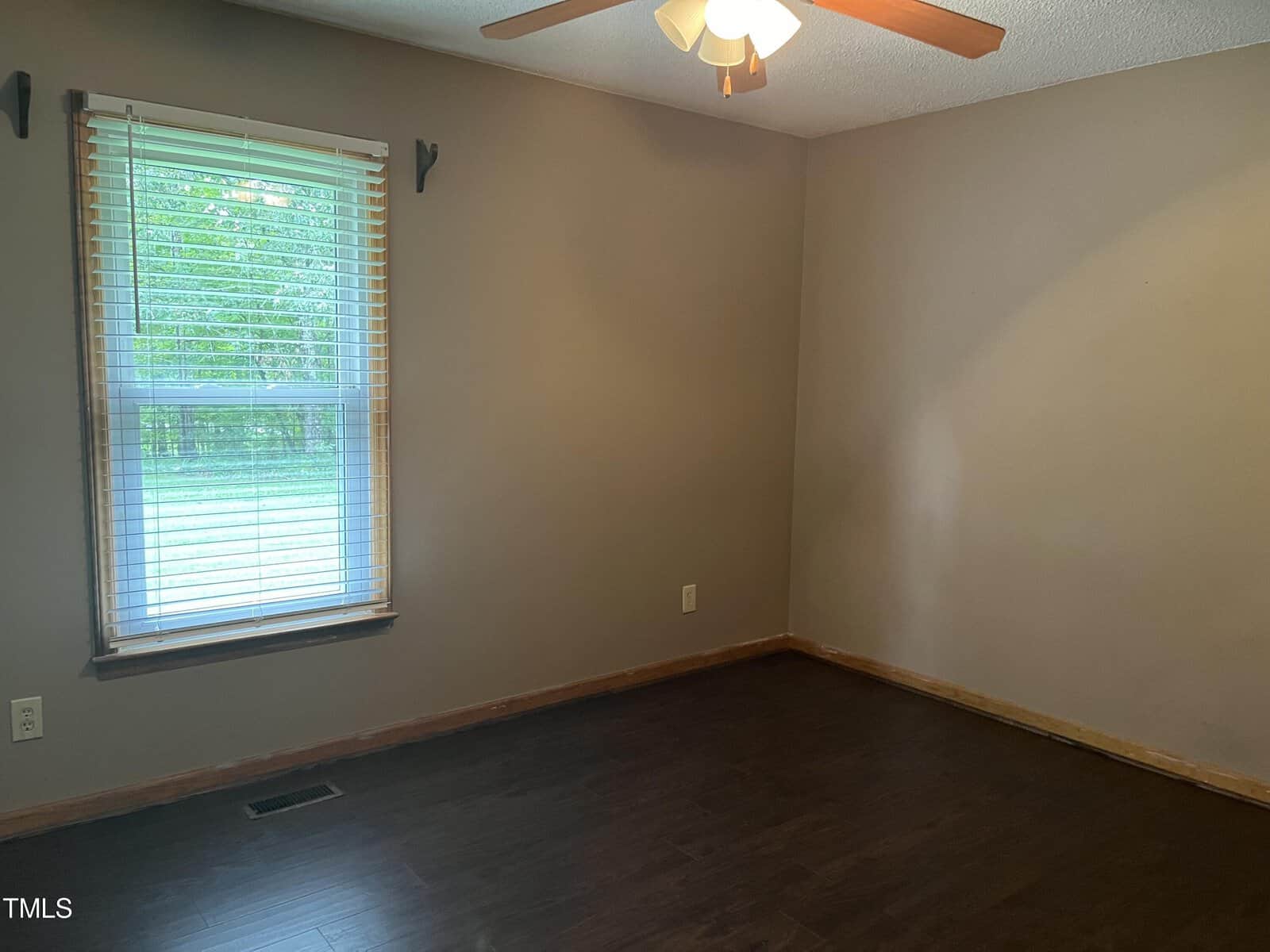 An empty room with beige walls and a dark wooden floor. There is a window with blinds on the left side, allowing natural light to enter. A ceiling fan with a light is installed above.