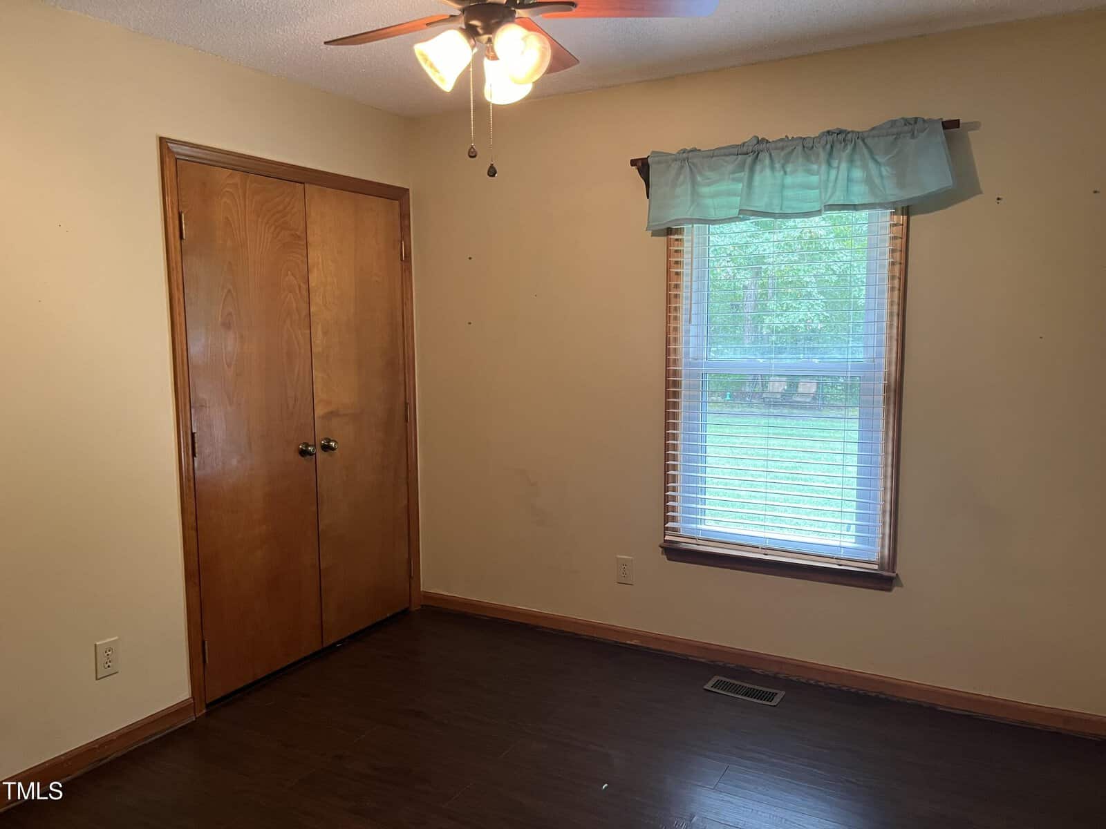 An empty room with light brown walls and dark wood floors. A ceiling fan with lights is above. There's a wooden closet door on the left and a window with blinds and a blue valance on the right, showing a green outdoor scene.