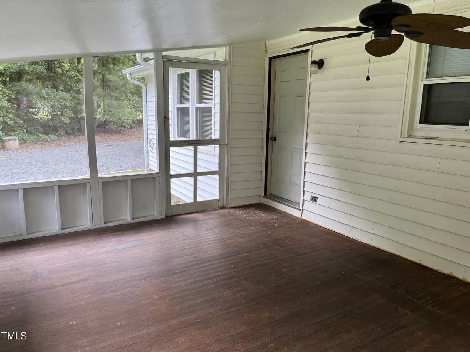A screened-in porch with white walls and a wooden floor, featuring a ceiling fan. The area is empty, and a doorway leads inside the house. Large windows provide a view of the wooded backyard and a gravel path outside.