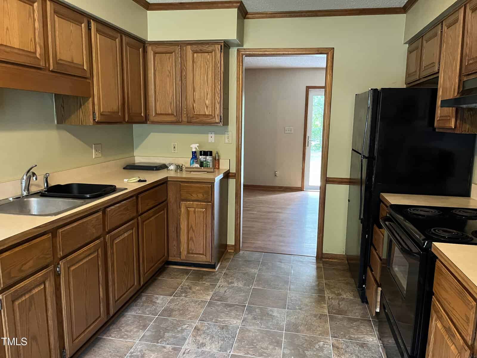 A kitchen with brown wooden cabinets, black appliances, and a tiled floor. The sink is on the left, and the stove is on the right. A doorway leads to a room with wooden flooring and a window in the background.