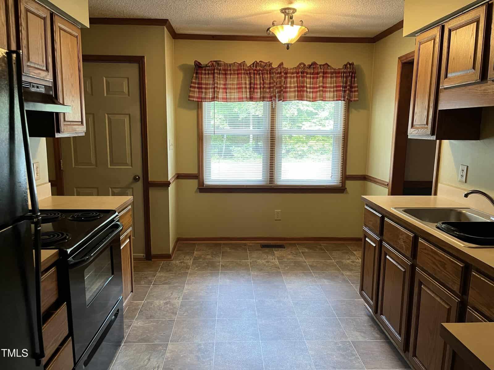 A kitchen with wooden cabinets and black appliances, including a stove and refrigerator. There's a large window with a red plaid valance, letting in natural light. The room has a tiled floor and a ceiling light fixture.