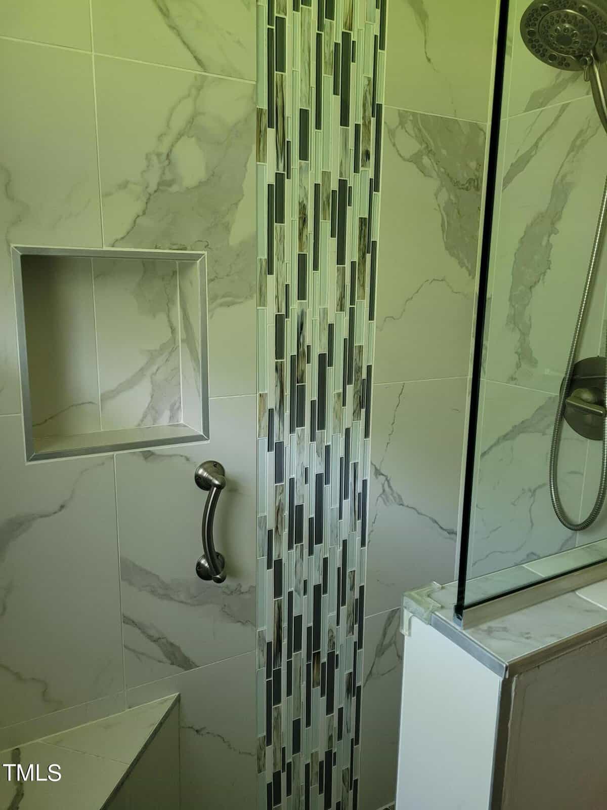 A modern shower with white and gray marble tile, featuring a vertical accent strip of smaller rectangular tiles. There's a built-in shelf niche, a metal grab bar, and a glass shower door. The showerhead is visible on the right side.