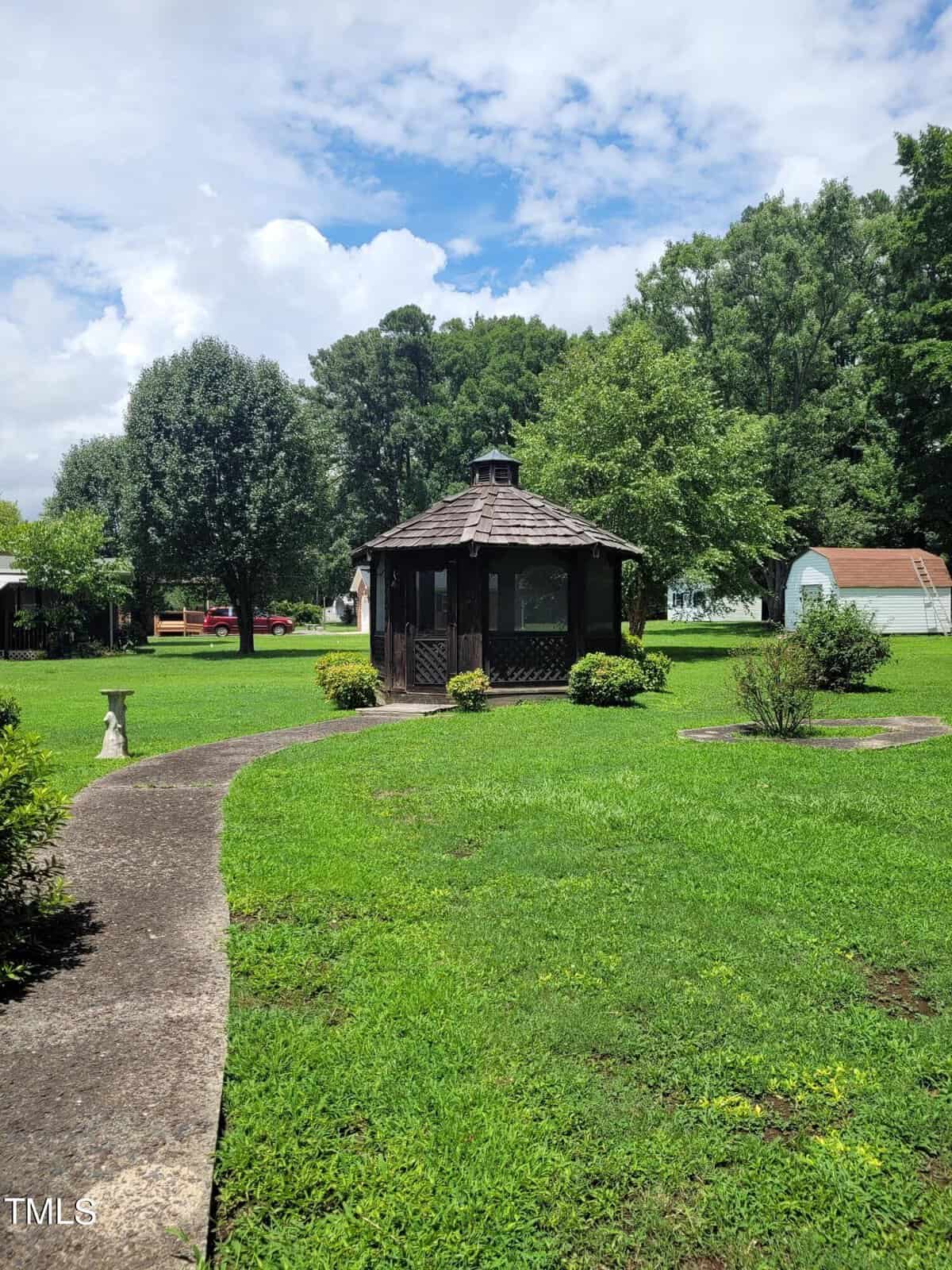 A garden scene with a circular gazebo at the center, surrounded by a well-maintained lawn. A curved concrete pathway leads to the gazebo, with trees, shrubs, and a shed in the background under a partly cloudy sky.
