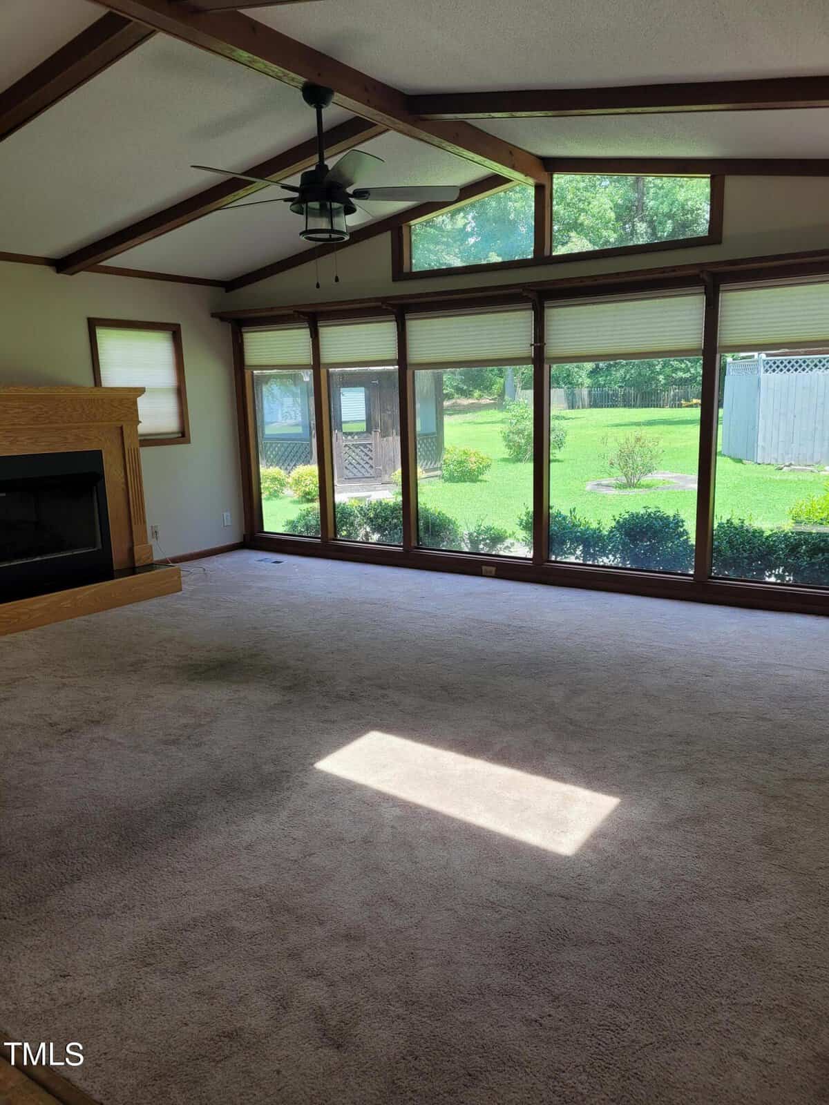 A spacious, empty living room with carpeted flooring, a wooden ceiling beam, and a ceiling fan. Large windows offer a view of a lush green backyard and garden. There's a fireplace on the left wall. Bright sunlight illuminates the room.