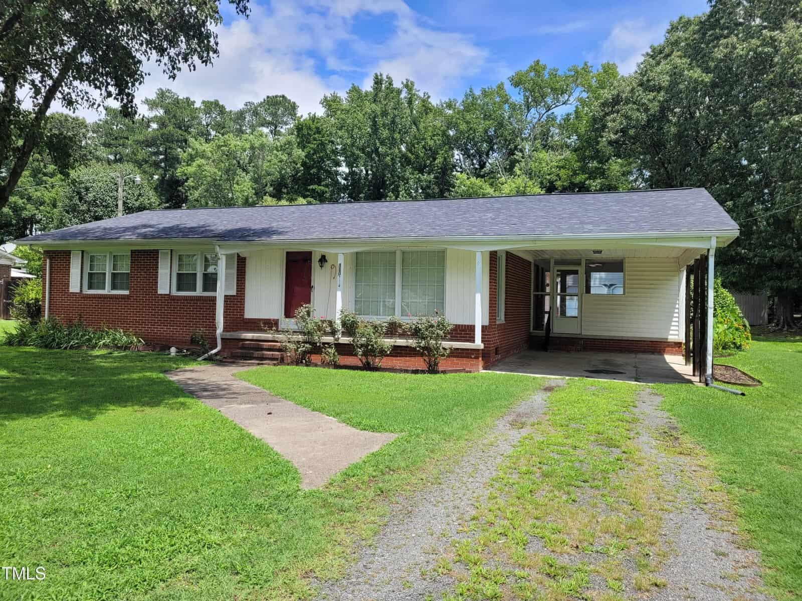 Located at 310 14th Street in Butner, this single-story brick house with white accents boasts a covered carport on the right. The dark roof shelters shrubs by the bay window, while a stone walkway gracefully leads to the iconic red front door, all framed by lush lawn and trees.