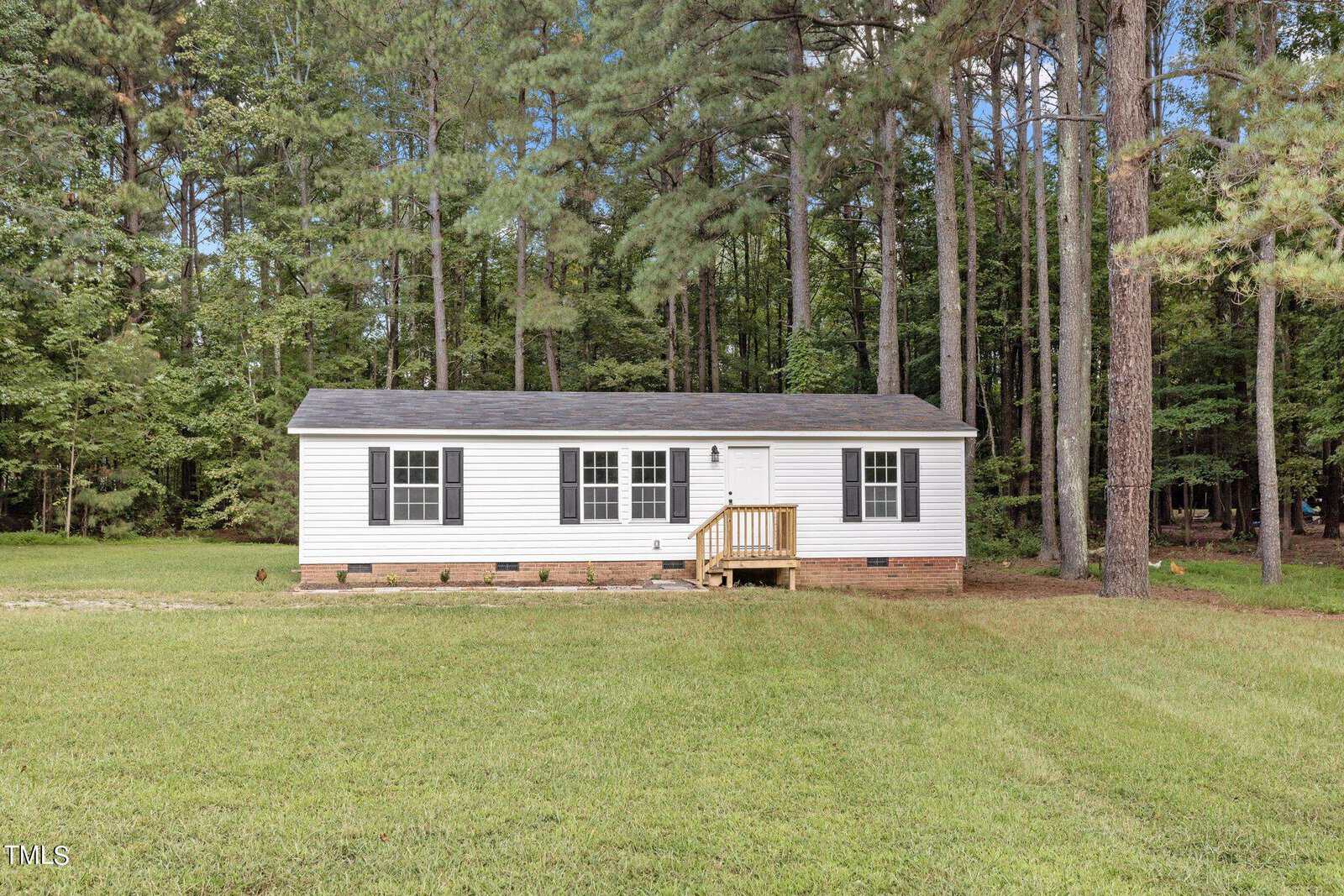 A small white house with a dark roof and black shutters is set against a background of tall pine trees. There is a wooden porch at the entrance, and the house is surrounded by a grassy yard under a clear blue sky.