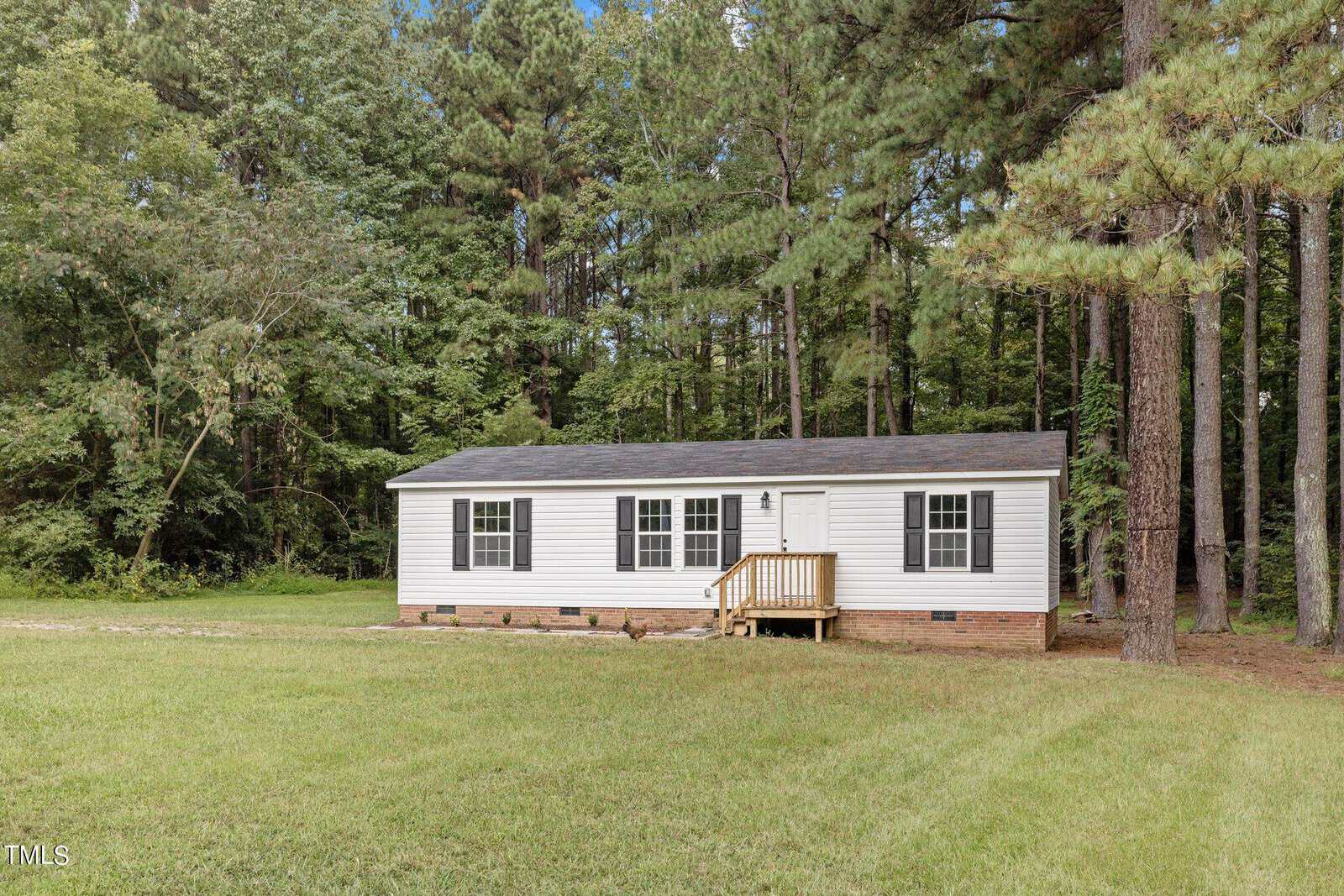 A small, white single-story house with black shutters is nestled among tall pine trees. The house has a wooden porch and sits on a large grassy lawn, surrounded by a wooded area.