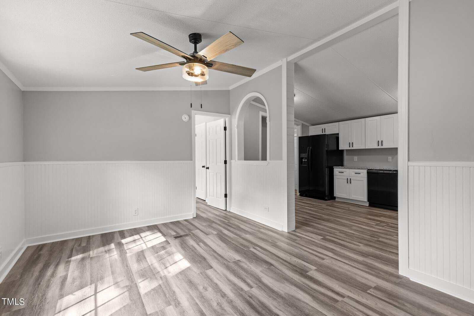 A spacious, modern kitchen and dining area with light wood flooring, gray walls, and white wainscoting. It features white cabinets, black appliances, and a ceiling fan with wooden blades. An arched opening leads to a different section of the home.