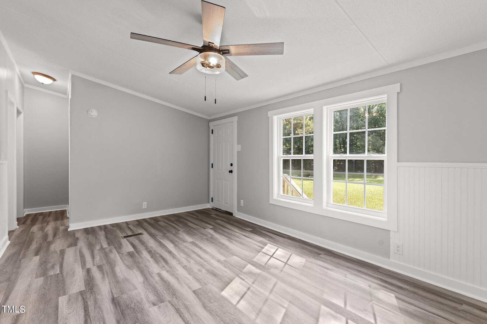 A bright room with light gray walls and vinyl wood flooring. A ceiling fan with a light fixture is centered on the ceiling. Two large windows allow natural light, with a view of a green lawn. A white paneled door leads to a hallway.