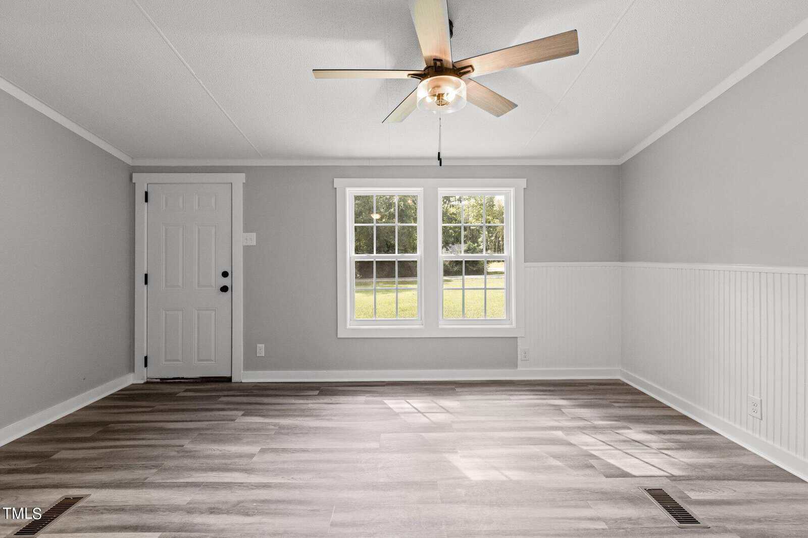 An empty room with gray walls and wood flooring features a ceiling fan with a light. A large window lets in natural light, overlooking a grassy yard. A white-paneled door is on the left. The wainscoting adds a classic touch to the walls.