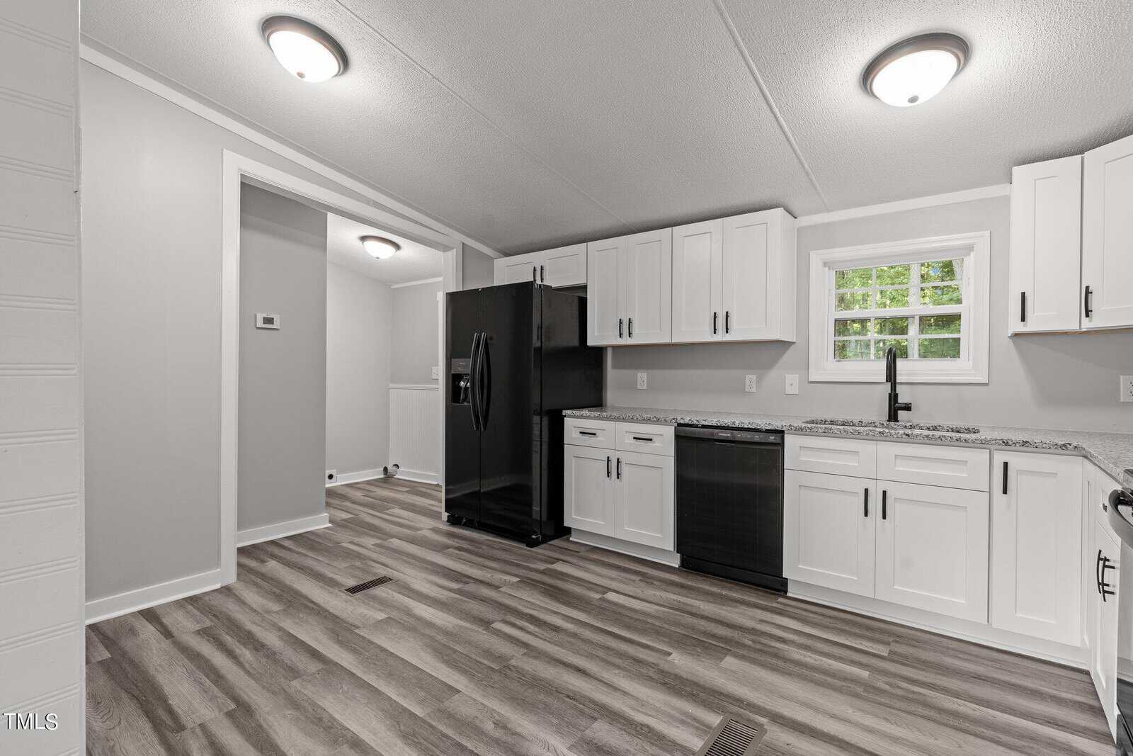 A modern kitchen with white cabinets, black appliances, and laminate wood flooring. The room features a large black refrigerator, black dishwasher, and multiple ceiling lights. A window above the sink offers natural light.