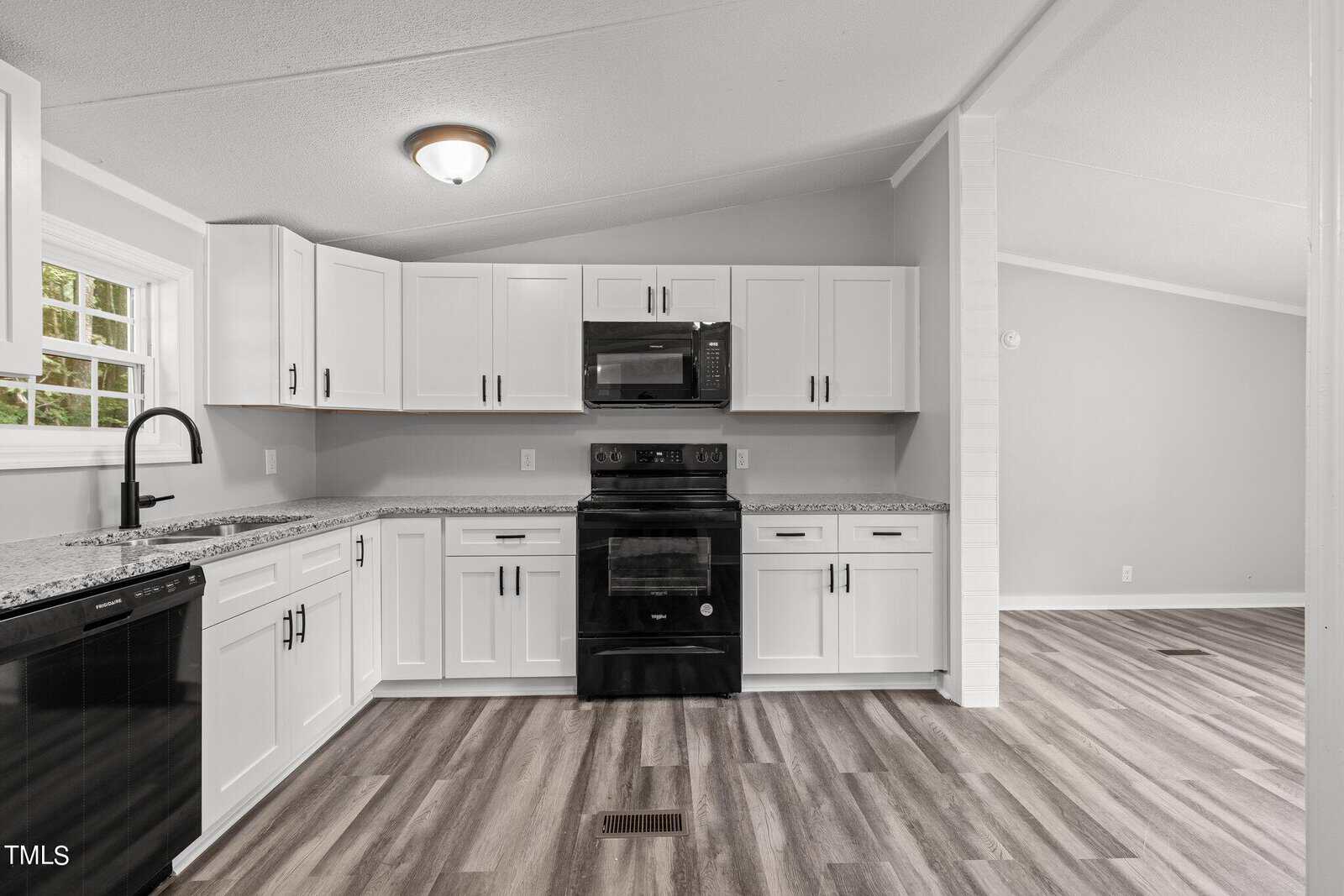 A modern kitchen with white cabinets and granite countertops. The appliances are black, and there's a window above the single basin sink. The floor is wooden, and the room is well-lit, with neutral gray walls.