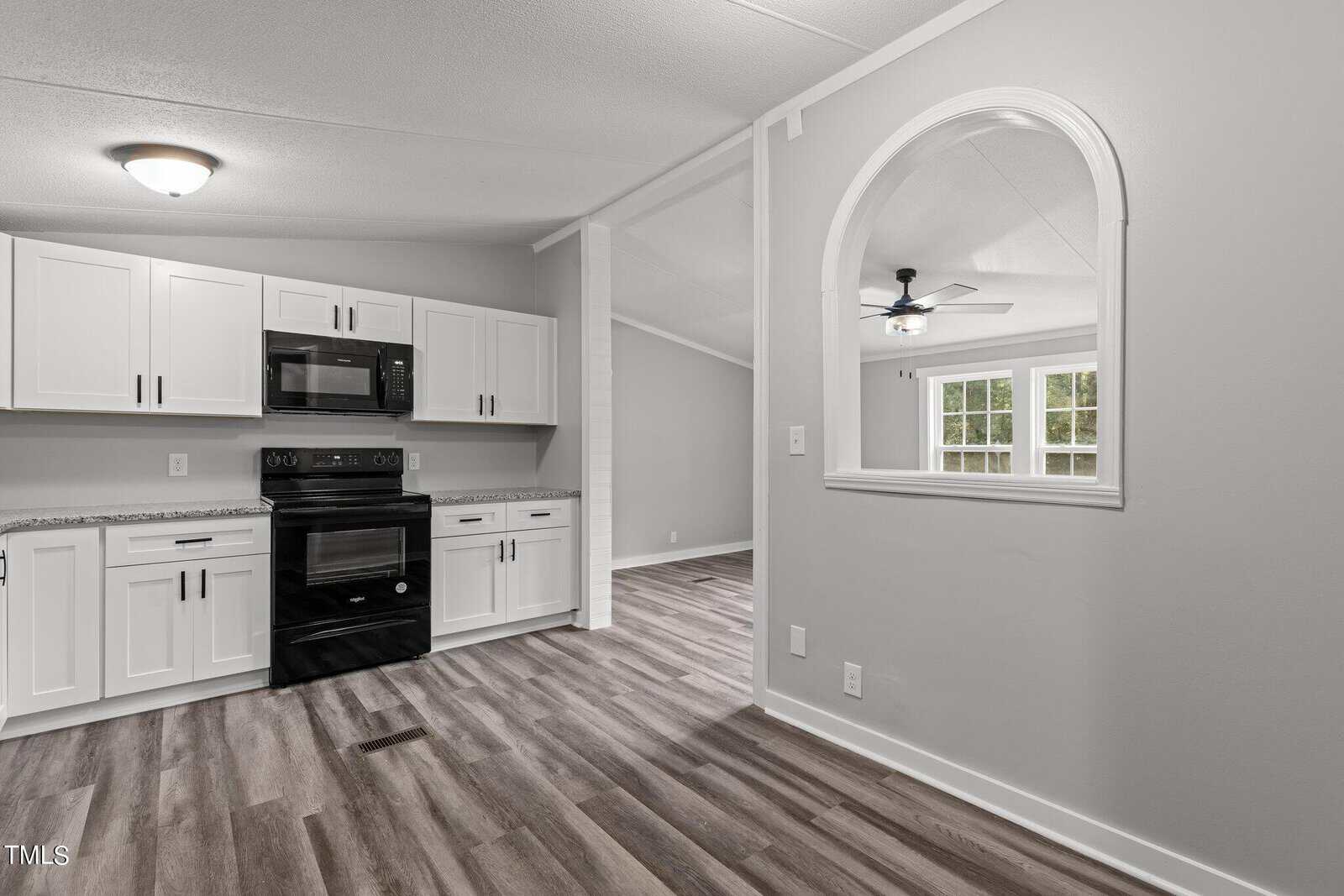 Modern kitchen with white cabinets, black appliances, and gray granite countertops. There's a pass-through archway to the adjacent room. The floor features wood-like vinyl planks, and the walls are painted light gray.