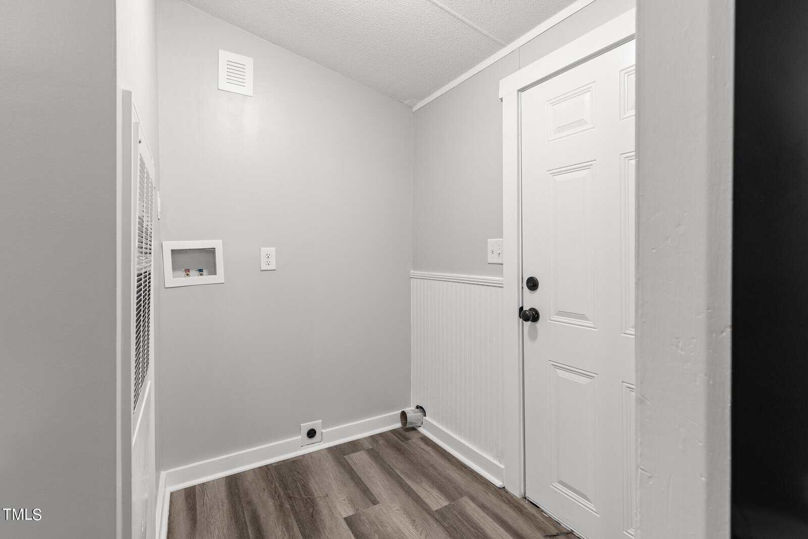 A small, empty room with light gray walls and a white door. The floor is covered with wood-like vinyl planks. There's a small square wall vent and a power outlet on one wall. The room is brightly lit, giving it a clean and minimalistic look.