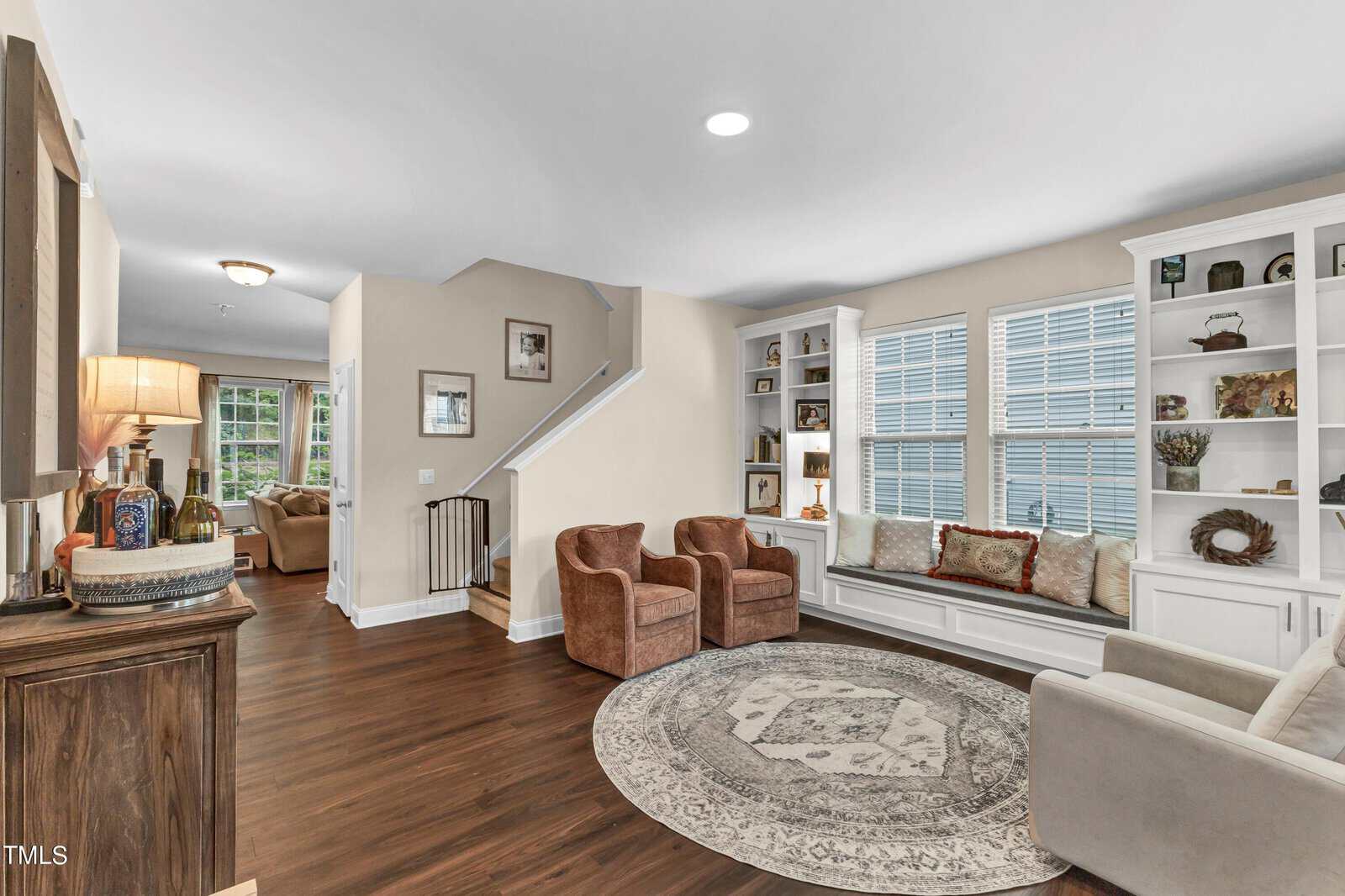 A living room with hardwood floors, built-in shelves and window seat, three armchairs around a circular rug, and a view of a hallway with stairs leading up. Large windows line one wall, letting in natural light.