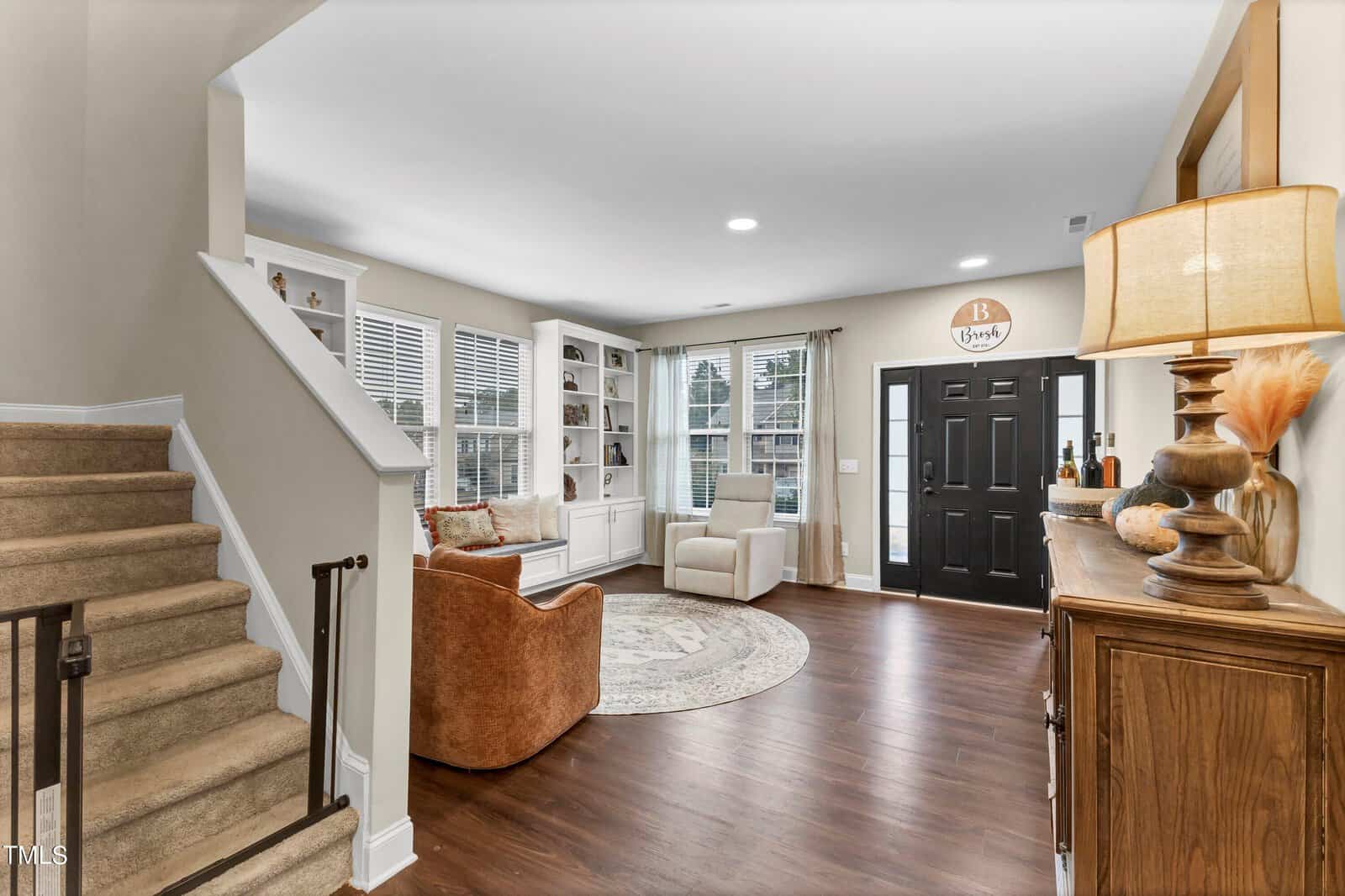 A living room with wood flooring, beige walls, a brown armchair, a round rug, built-in white shelves, a black front door, a staircase with carpeted steps, and a wooden sideboard with a lamp and decor.