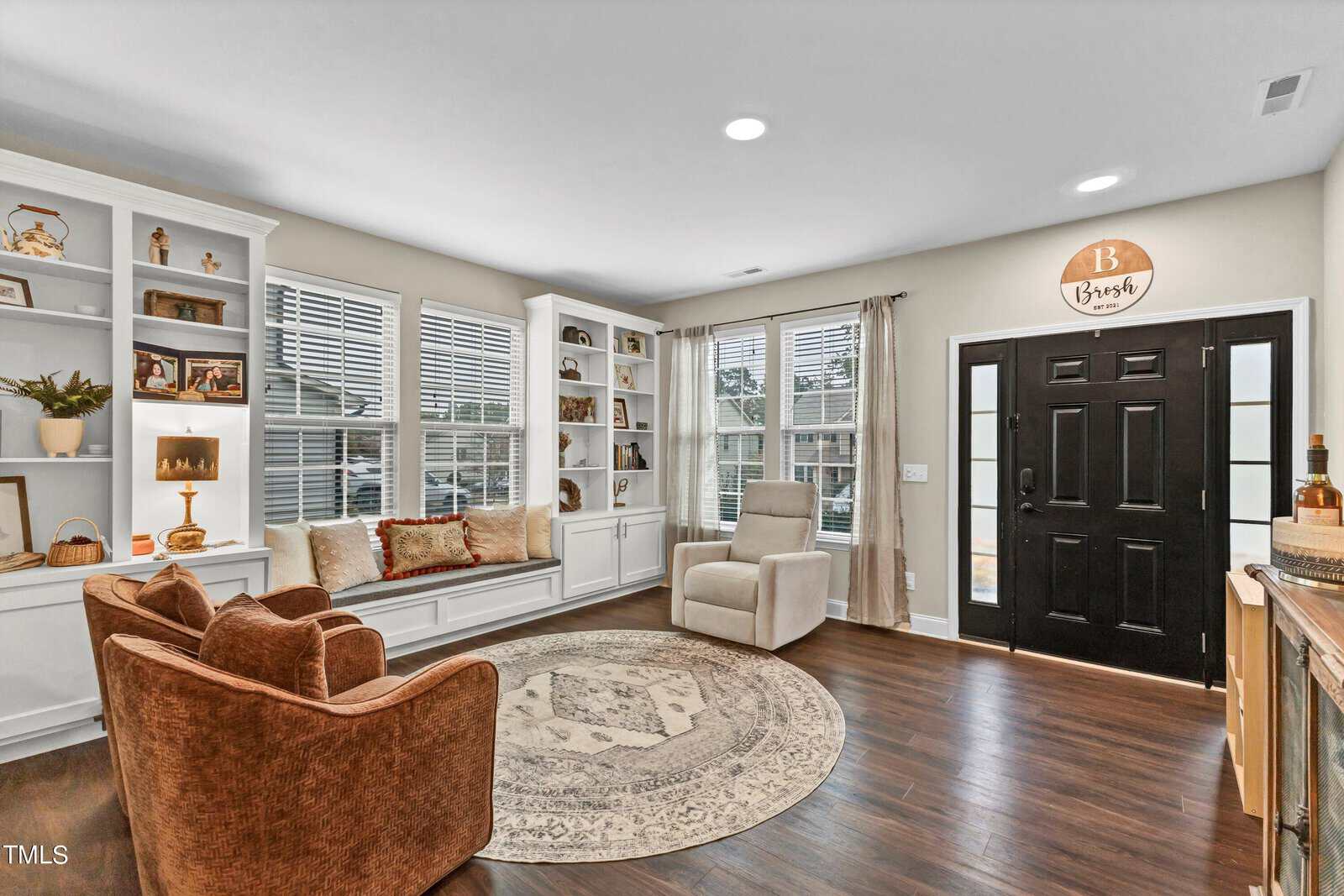 A bright living room with large windows, a black front door, built-in white shelves, a window seat with cushions, an armchair, a round rug, and wood floors. Decorative items are displayed on the shelves.