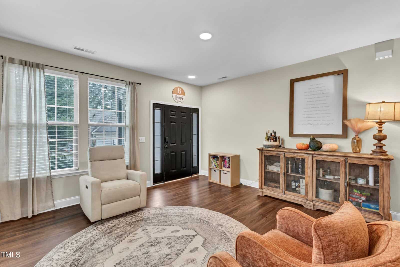 A living room with a beige recliner, an orange armchair, a round rug, wood flooring, a glass-front cabinet with decor items, a lamp, and a window with sheer curtains next to a black front door.