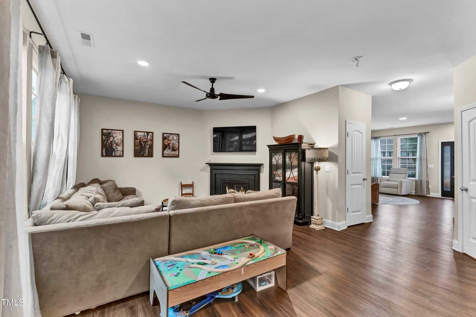 A cozy living room with a sectional sofa, wall art, TV above a fireplace, and children’s play table. Hardwood floors, neutral walls, and an adjacent seating area with large windows complete the space.