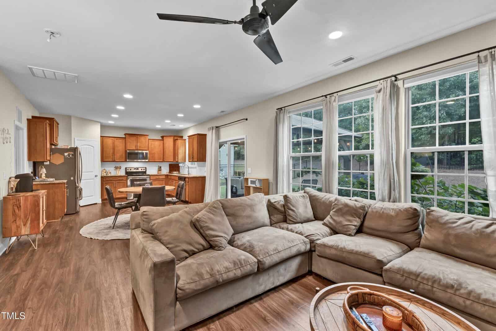 A spacious living room with a large brown sectional sofa, wooden floors, a ceiling fan, and large windows. The open layout leads to a kitchen with wood cabinets and stainless steel appliances.