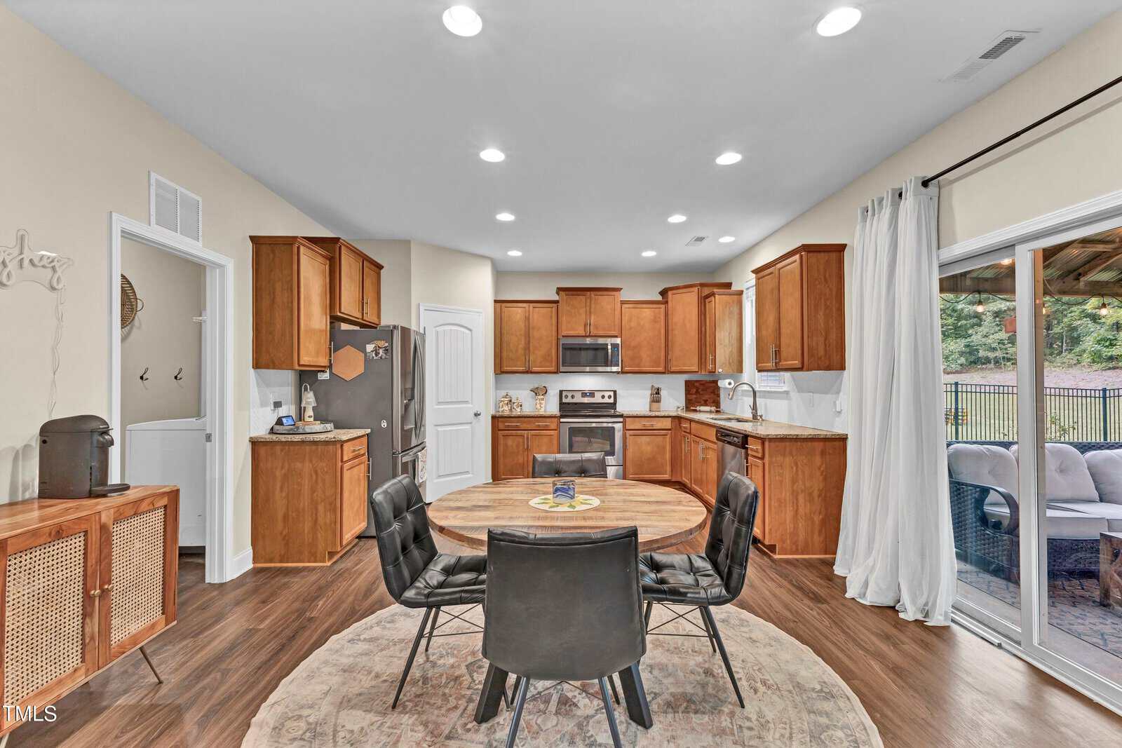 A modern kitchen with wooden cabinets, stainless steel appliances, and a round wooden dining table with four black chairs on a circular rug. Sliding glass doors open to an outdoor patio area.