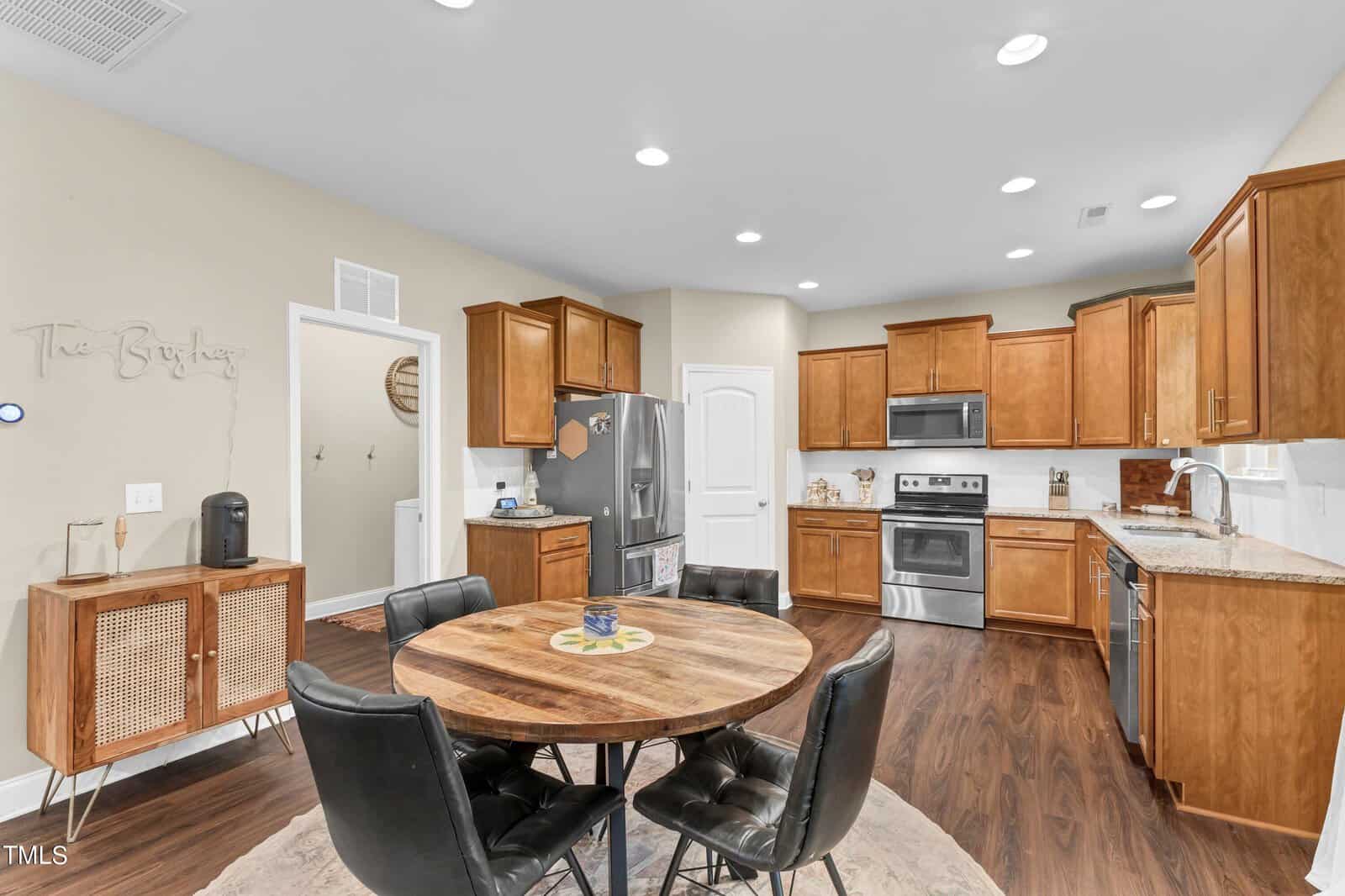 A modern kitchen and dining area with wooden cabinets, stainless steel appliances, a round wooden dining table with four black chairs, and wood-look flooring under bright recessed lighting.