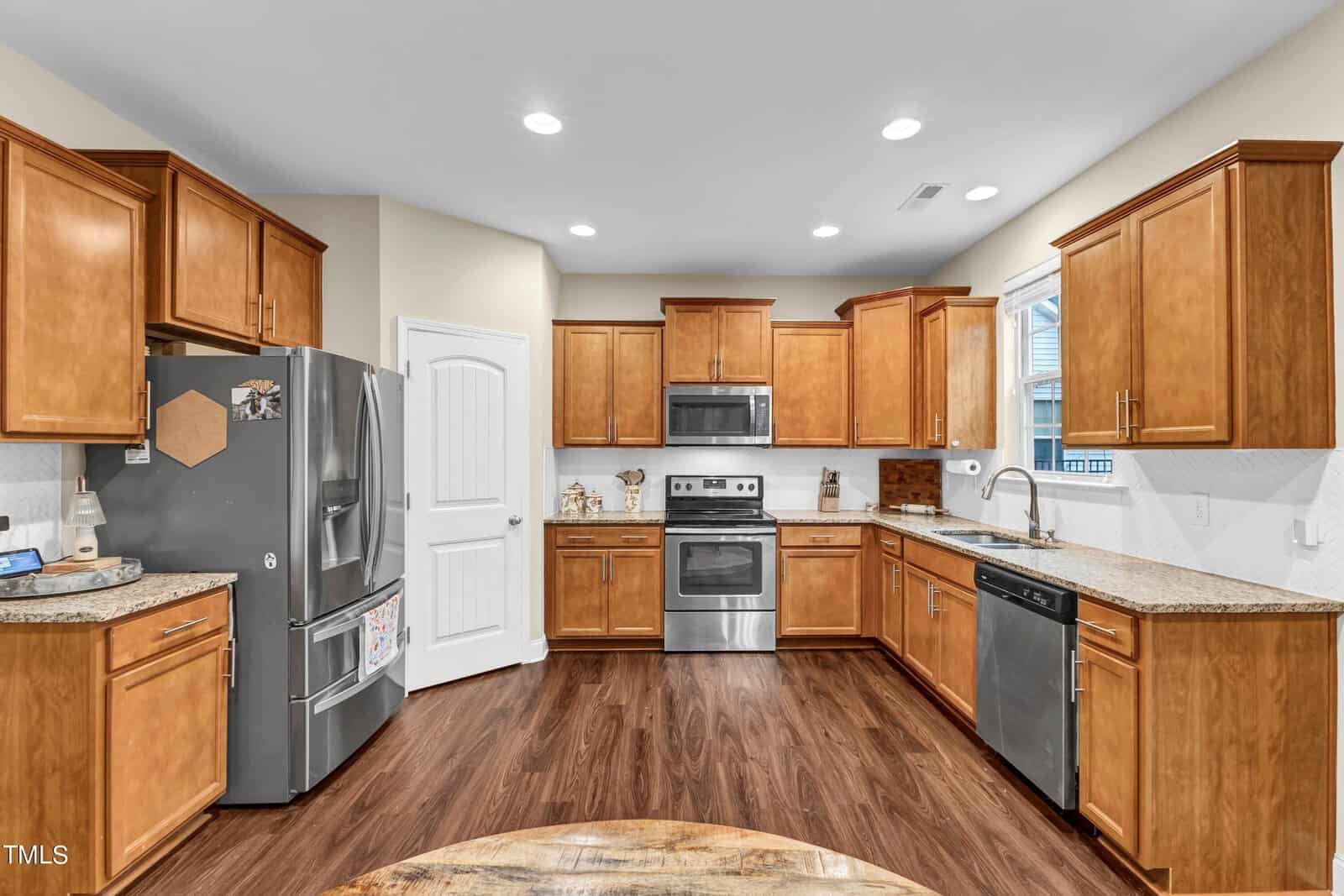 Modern kitchen with wood cabinets, stainless steel appliances including a refrigerator, oven, and microwave, granite countertops, and wood flooring. The space is well-lit with recessed lighting and has a tidy appearance.