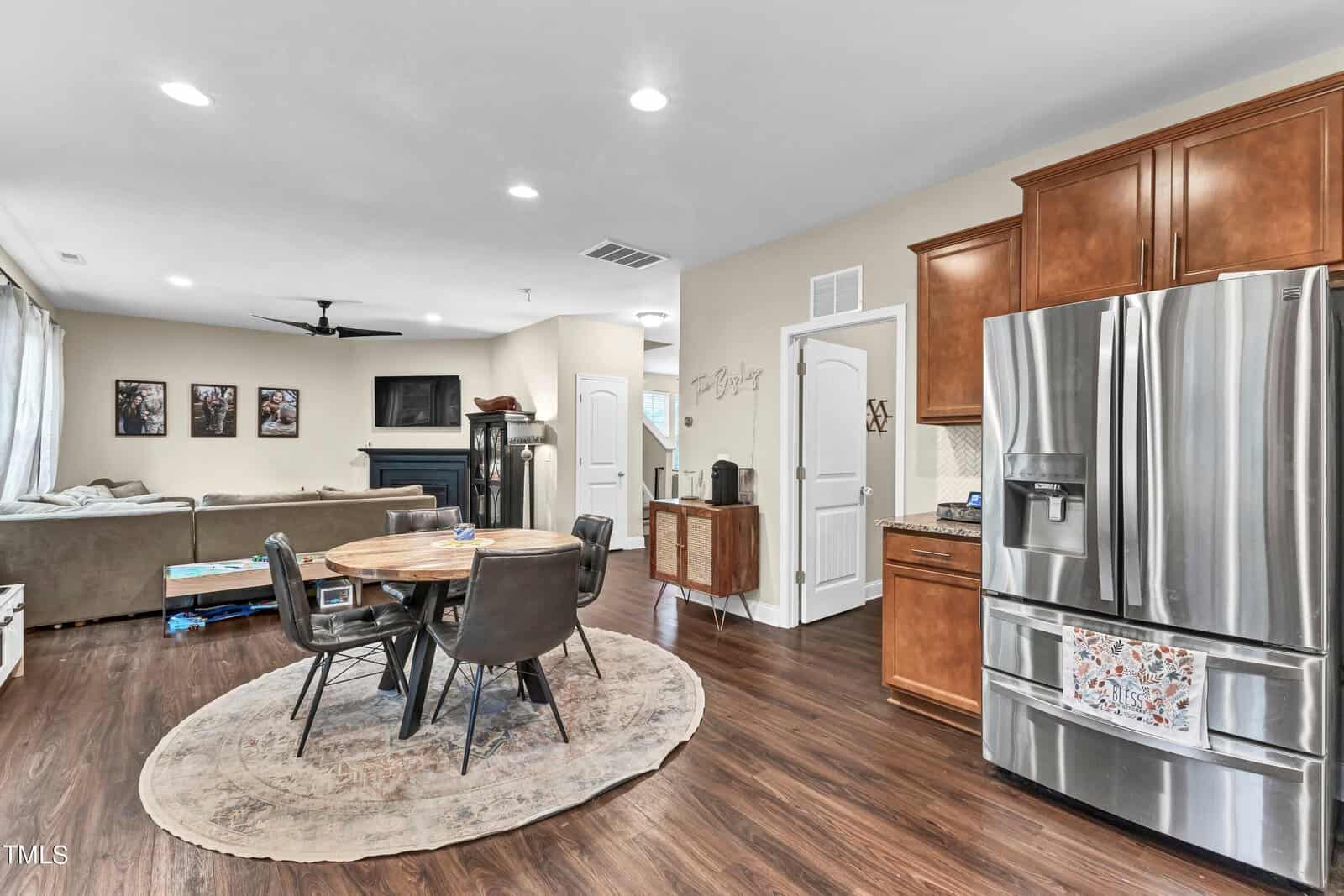 Open-concept kitchen and living area with stainless steel refrigerator, round dining table with four chairs on a rug, brown cabinets, sectional sofa, wall-mounted TV, and hardwood floors.