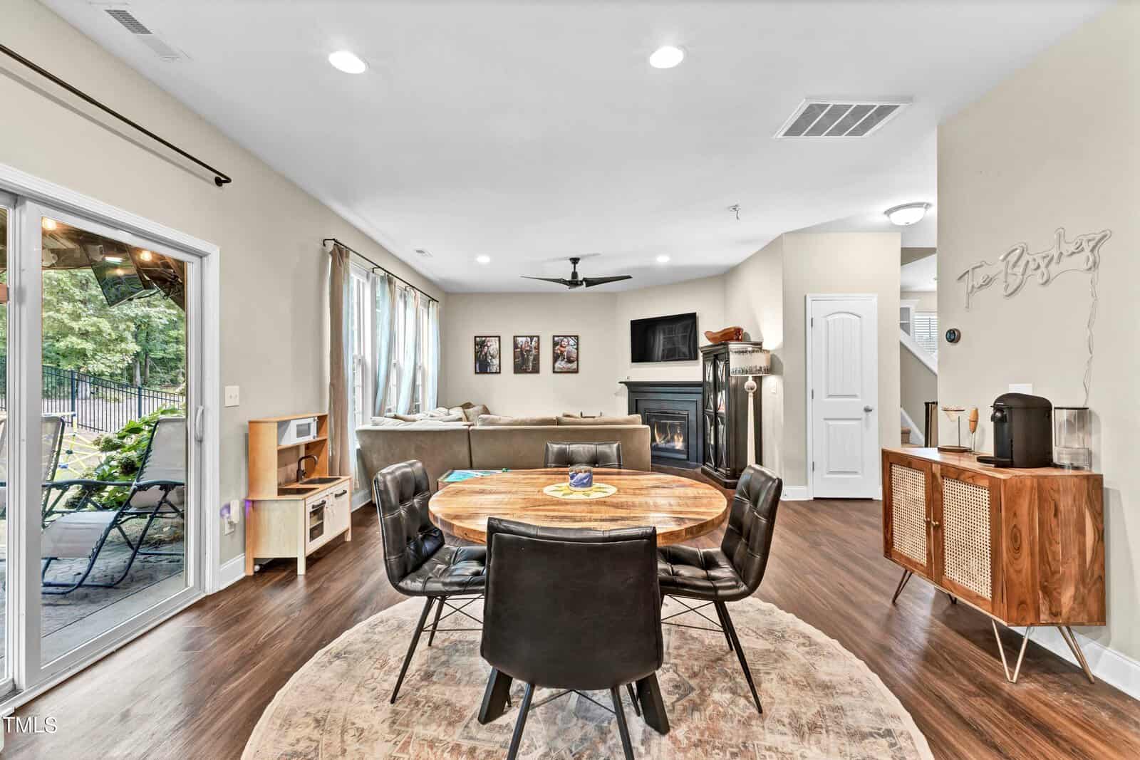 Open-plan living and dining area with a round wooden table and four black chairs on a circular rug; living space with a sectional sofa, TV, and fireplace; sliding glass door to a patio; wood floors and neutral walls.