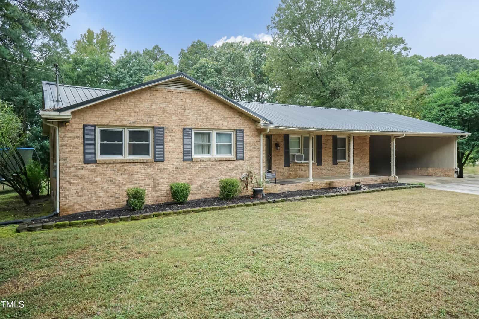 A single-story brick house with a metal roof, featuring a covered front porch supported by four columns and a carport to the right. The yard is partially landscaped with shrubs and trees in the background. The sky is clear, and the overall setting is calm and suburban.