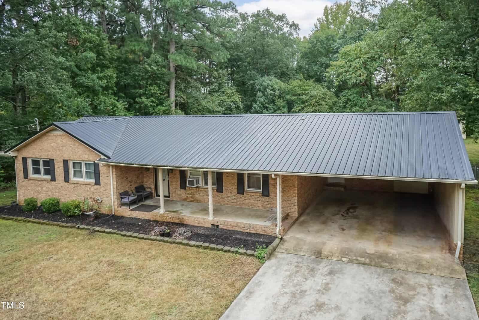 A single-story brick house with a metal roof, featuring a front porch with seating, a neat garden bed running along the front, and an attached carport on the right. The background shows tall trees surrounding the property. The driveway leads up to the carport area.