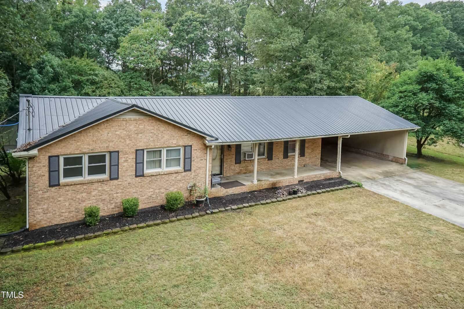 A single-story brick house with a large covered carport and a metal roof. The front yard has a mix of grass and small shrubs along the foundation. The background features tall, dense trees. The overall setting is suburban and peaceful.