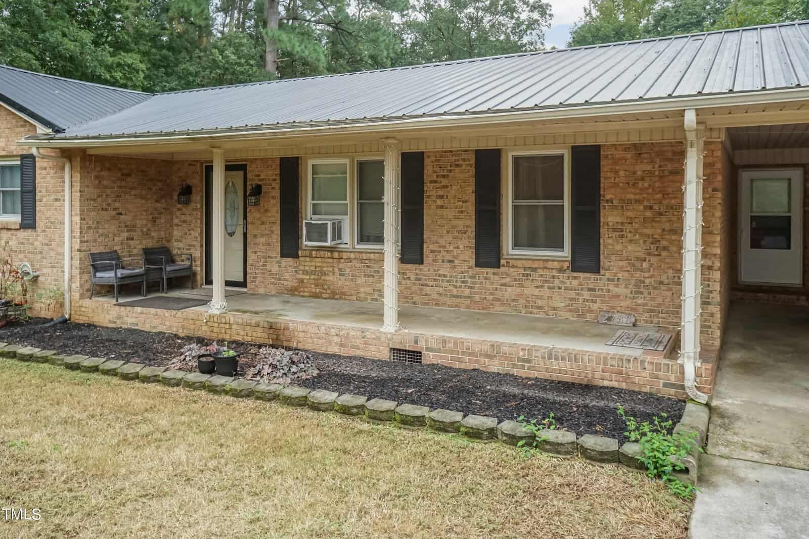 A single-story brick house with a metal roof features a covered front porch. There are two wicker chairs and a small table on the porch. Black window shutters and a window air conditioning unit are visible. The lawn is neatly trimmed, and there are garden beds along the porch.