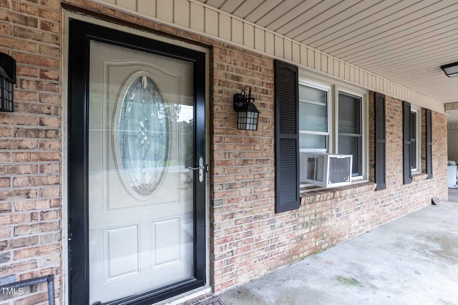 A residential brick house features a covered porch with a concrete floor. The front door is white with a decorative oval glass insert framed in black. Beside the door, there is a wall-mounted black lantern. Two windows with black shutters have window-mounted air conditioners.