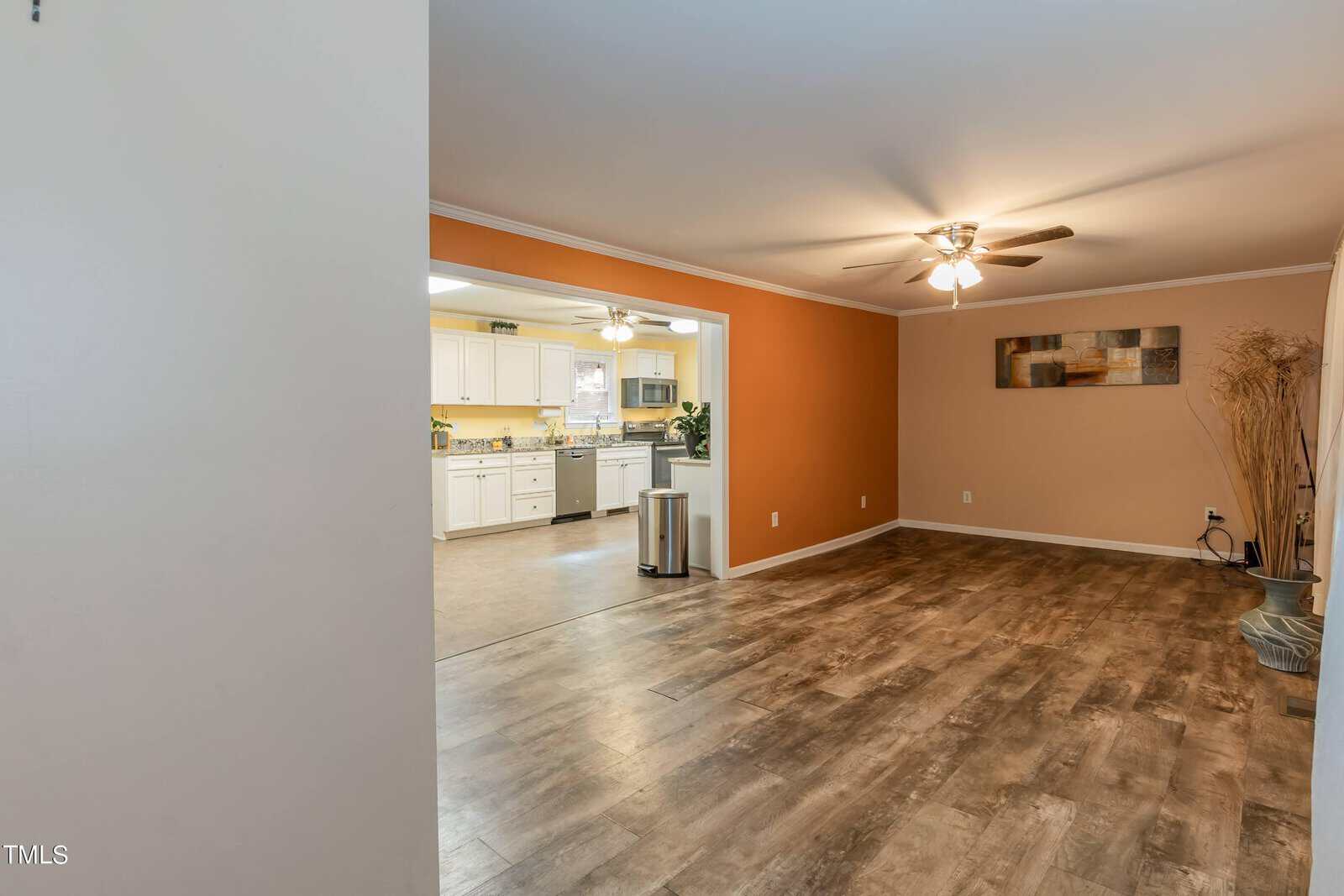 A spacious room with wood flooring and beige walls. The room has a ceiling fan with lights, and a piece of artwork hanging on the wall. The room opens into a kitchen with white cabinets and stainless steel appliances. The kitchen has yellow walls and tiled flooring.