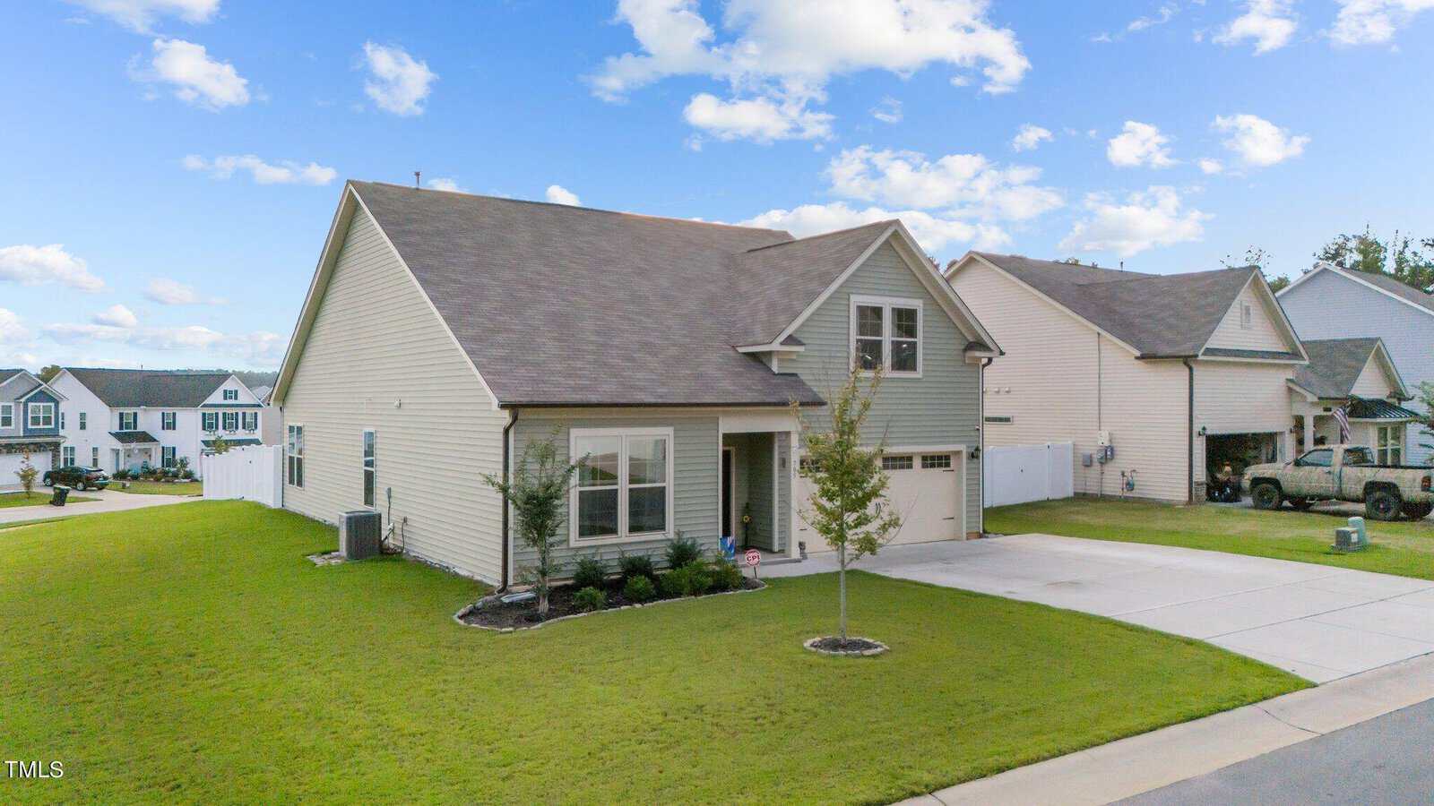 A light-colored, two-story suburban house with a gable roof and attached garage, surrounded by a green lawn and small trees, situated in a residential neighborhood under a partly cloudy sky.