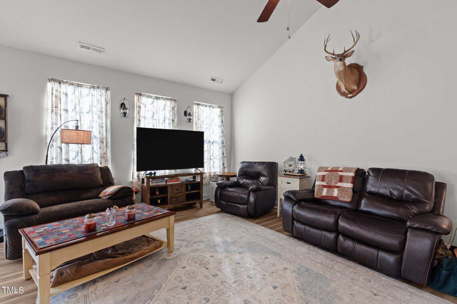 A living room with dark brown leather sofas and recliner, a wooden coffee table, a TV on a stand between two windows with curtains, and a mounted deer head on the wall. A ceiling fan is visible above.