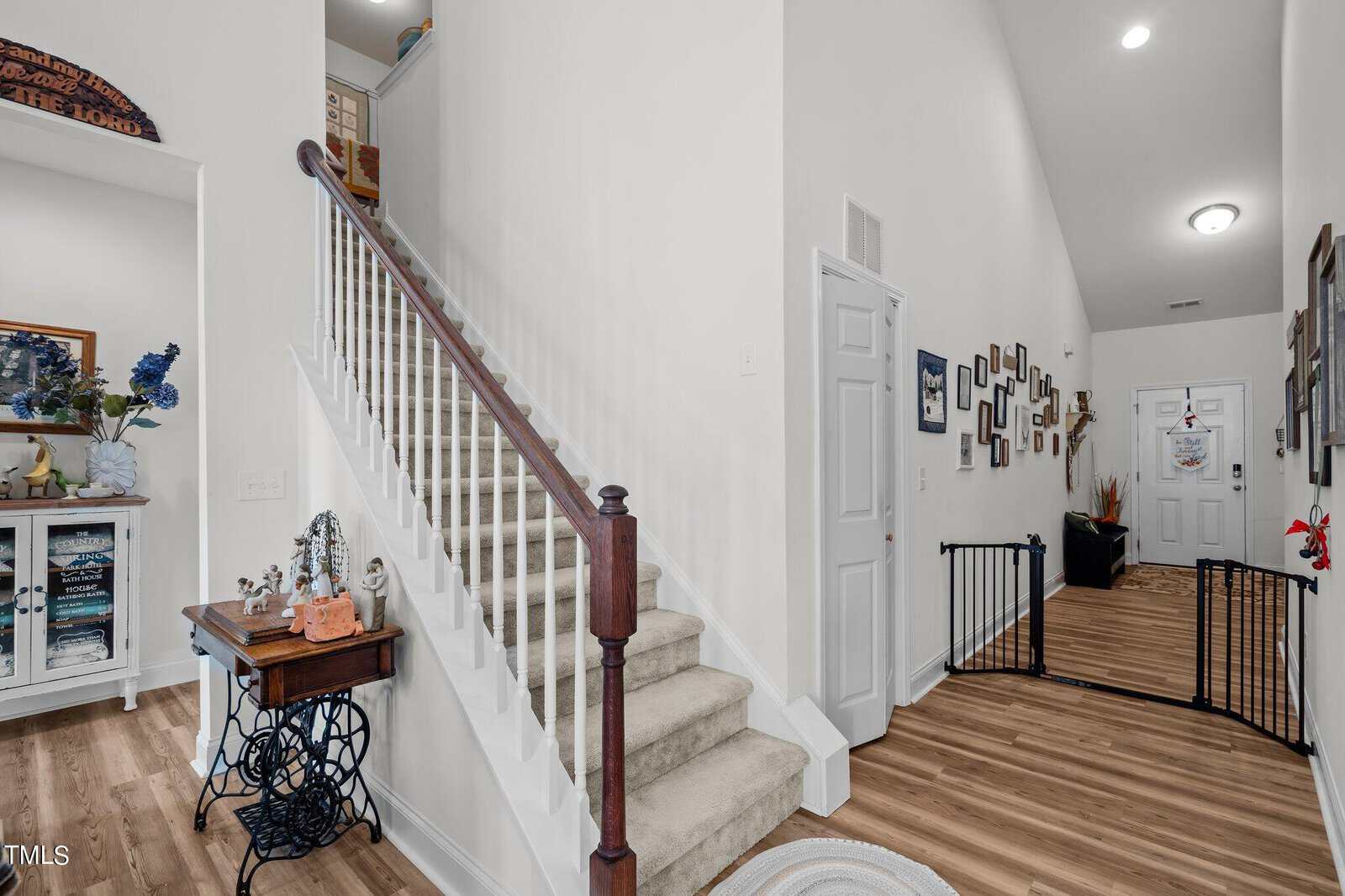 A carpeted staircase with a dark wooden handrail leads to an upper floor. The hallway features wood flooring, a white cabinet with decor, a gallery wall of photos, and a child safety gate near the front door.