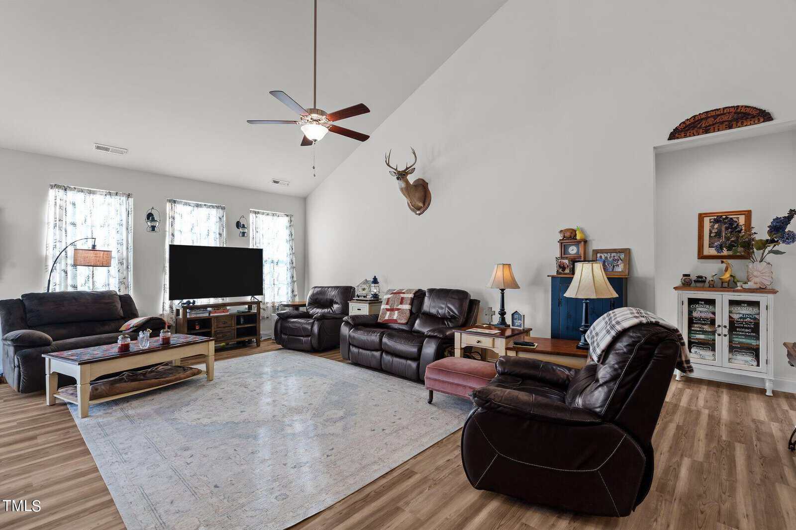 A spacious living room with brown leather recliners, a large TV, a coffee table, light-colored area rug, wood flooring, two windows with sheer curtains, and a mounted deer head on a white wall.