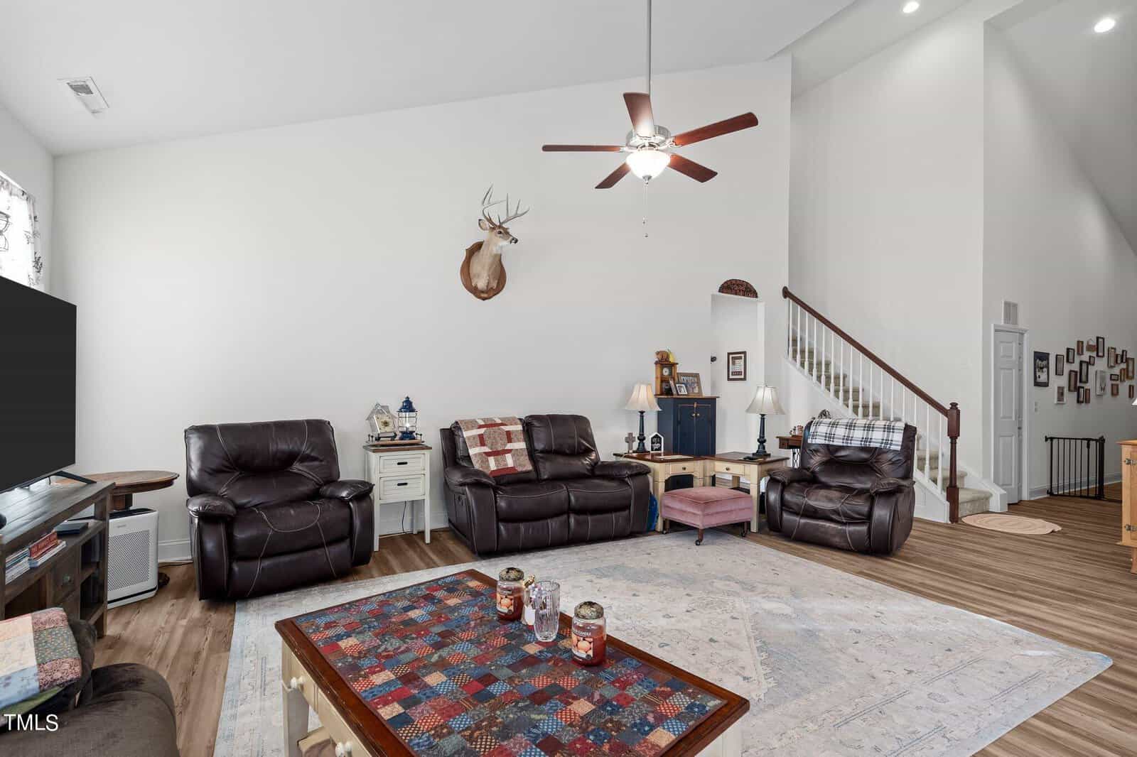 A living room with brown leather seating, a patterned coffee table, and a mounted deer head on a white wall. There's a ceiling fan, stairs to the right, and a mix of cabinets and side tables.
