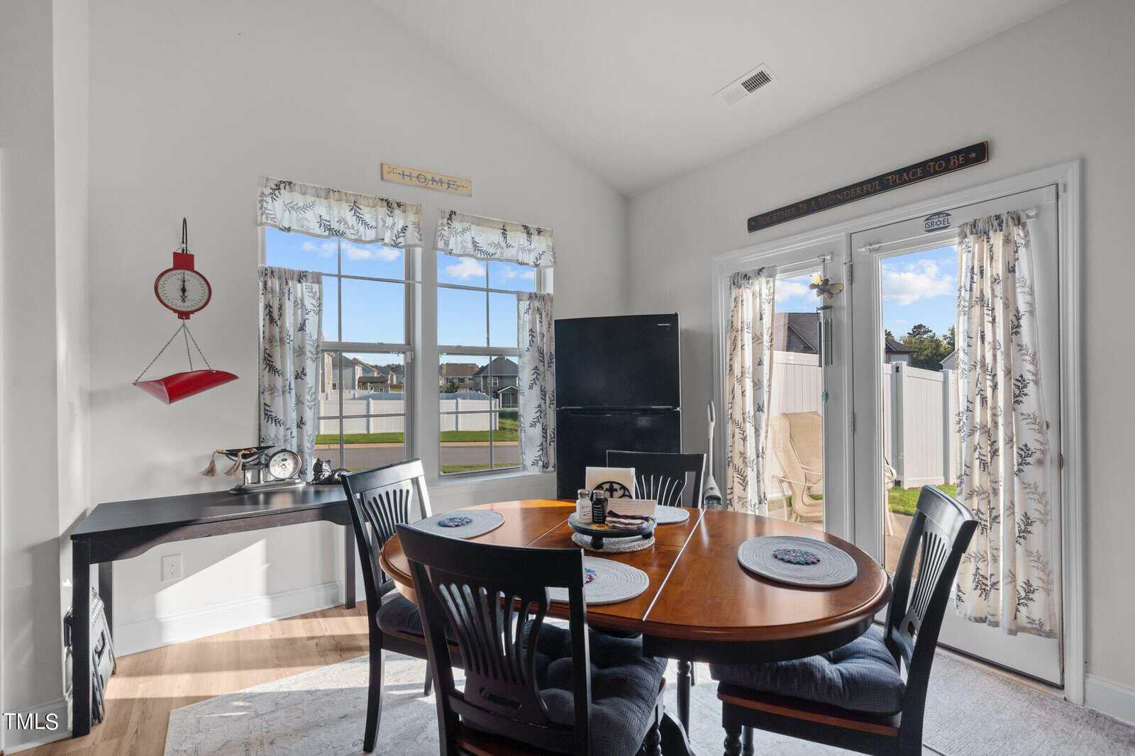 Bright dining area with a round wooden table, four black chairs, black fridge, wall clock, and decor. Large windows and glass doors let in sunlight, revealing a fenced backyard and suburban houses outside.