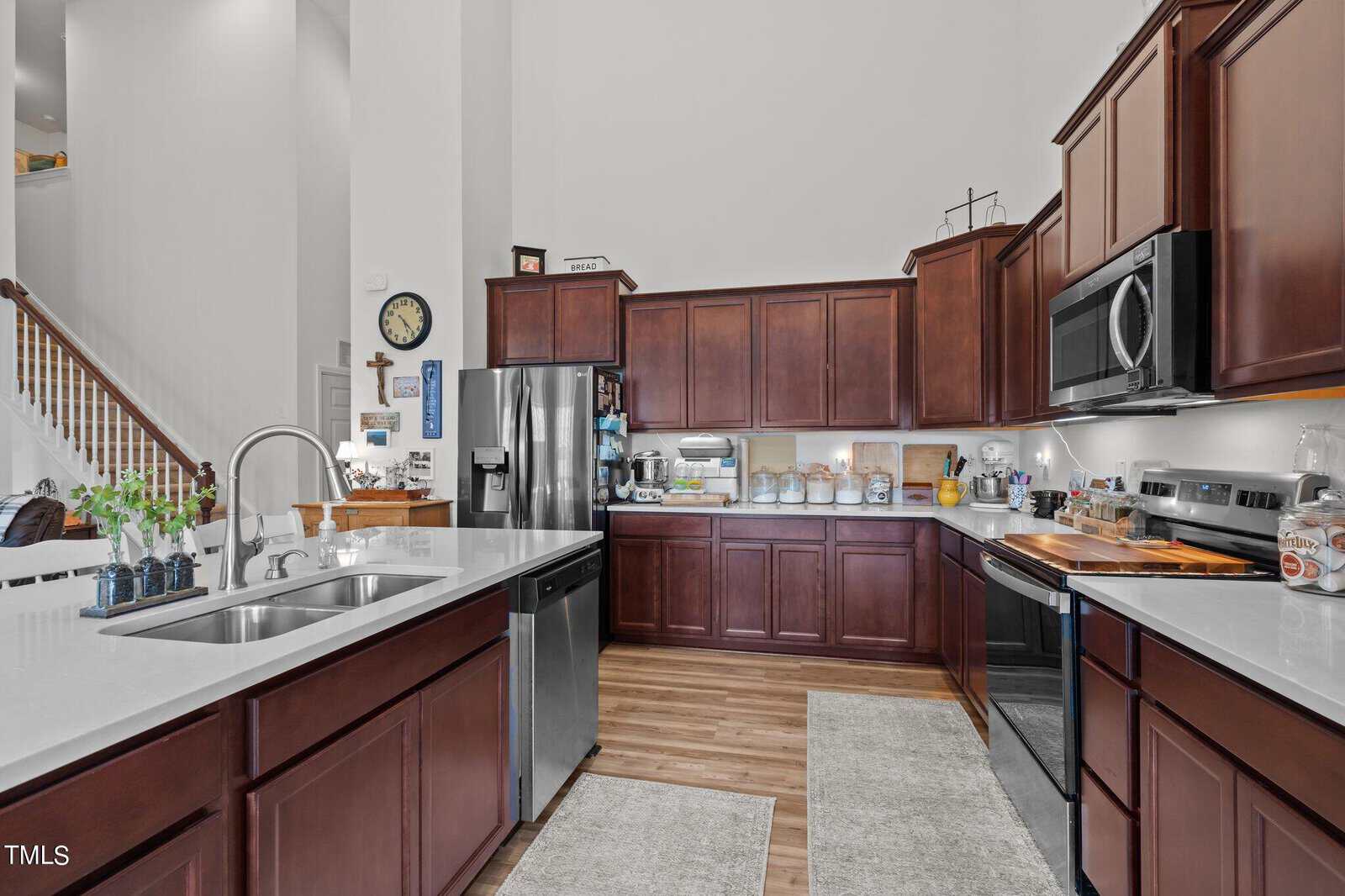 A modern kitchen with dark wood cabinets, stainless steel appliances, white countertops, a double sink on an island, and light wood flooring. Various small appliances and kitchen items are on the countertops.