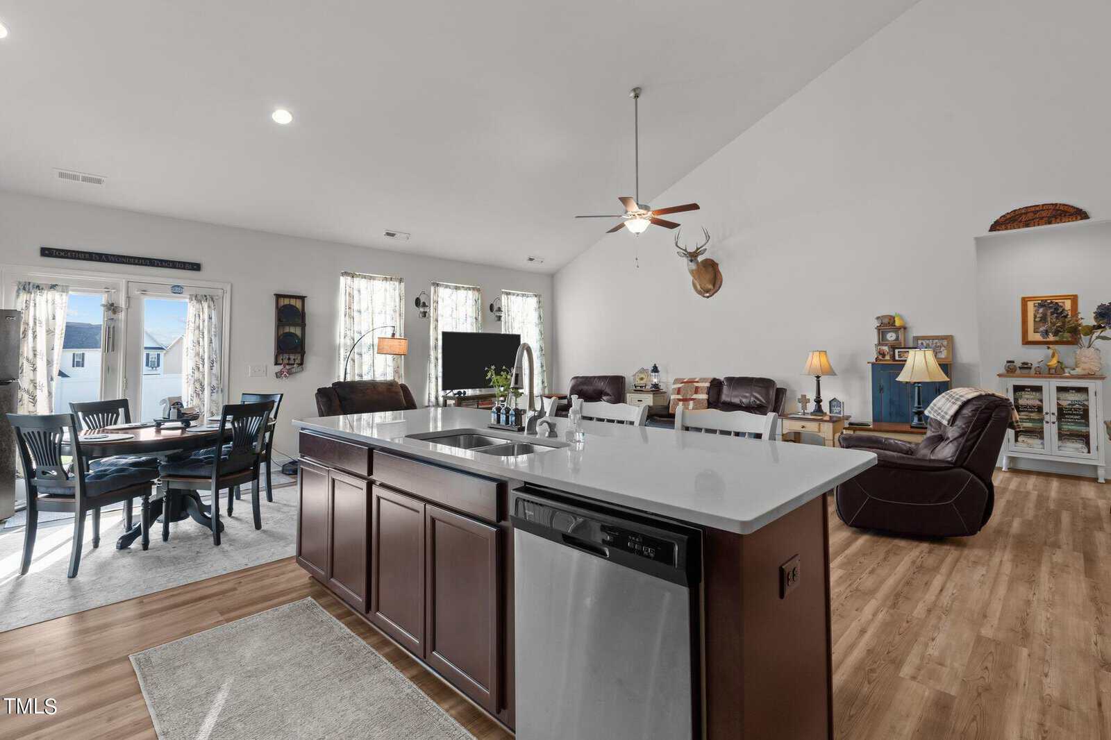 Open-concept living space with a kitchen island in the foreground, stainless steel dishwasher, dining table to the left, and living area with sofas, armchairs, TV, and decor on the walls and shelves.