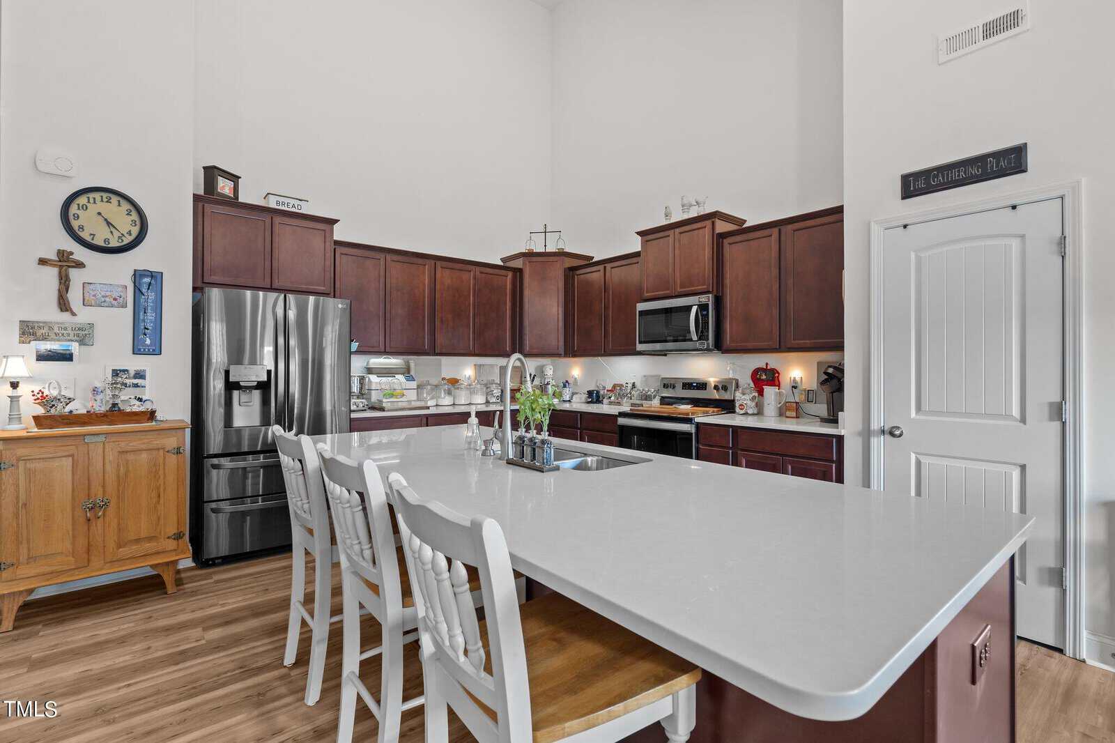 Modern kitchen with dark wood cabinets, stainless steel appliances, a white island with sink, and four white chairs. Wooden floors, wall decorations, and various items on counters create a cozy, lived-in feel.