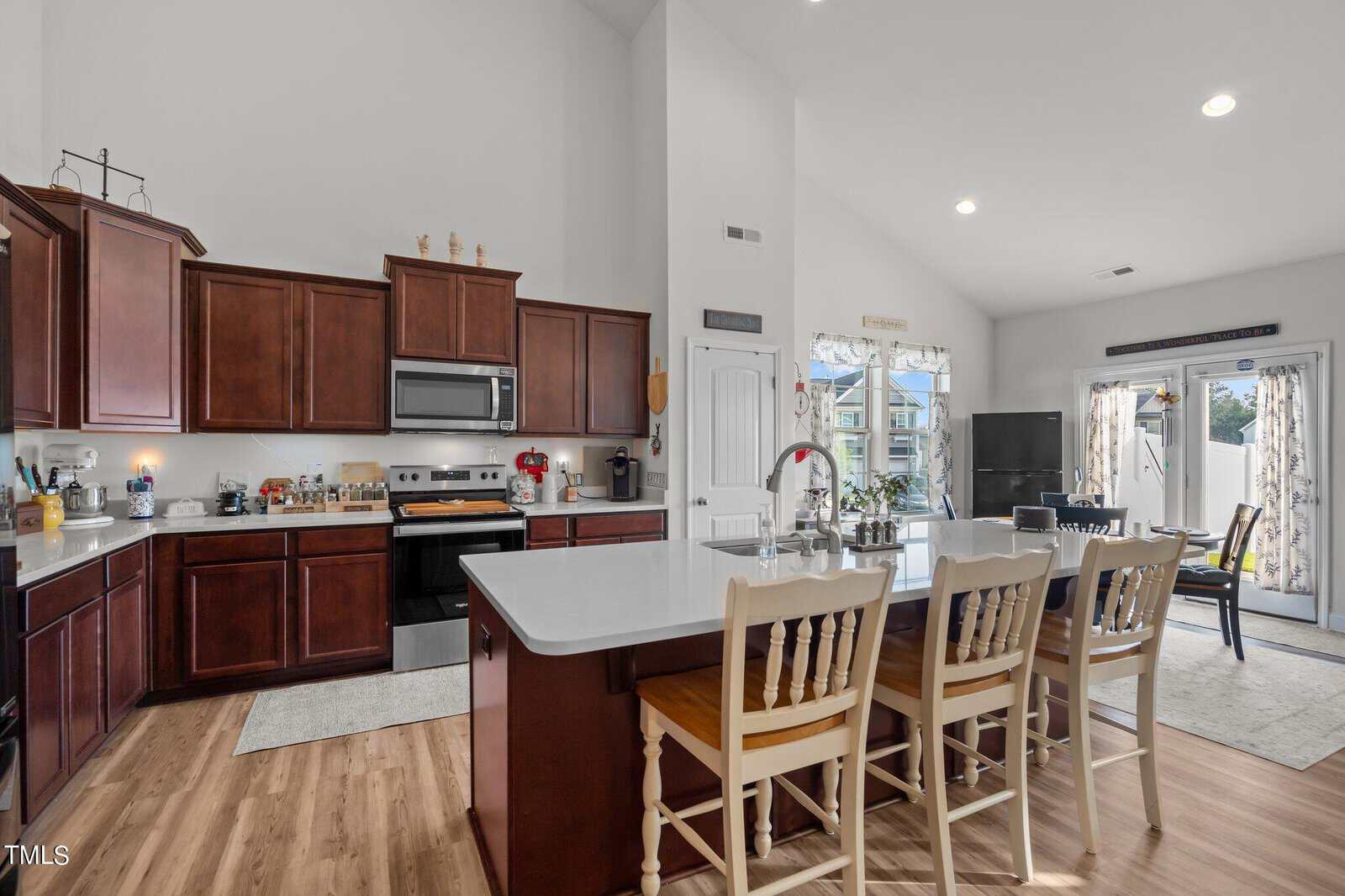 A spacious kitchen with dark wood cabinets, stainless steel appliances, a large white island with a sink, and three wooden chairs. The room is well-lit with natural light from windows and a glass door.