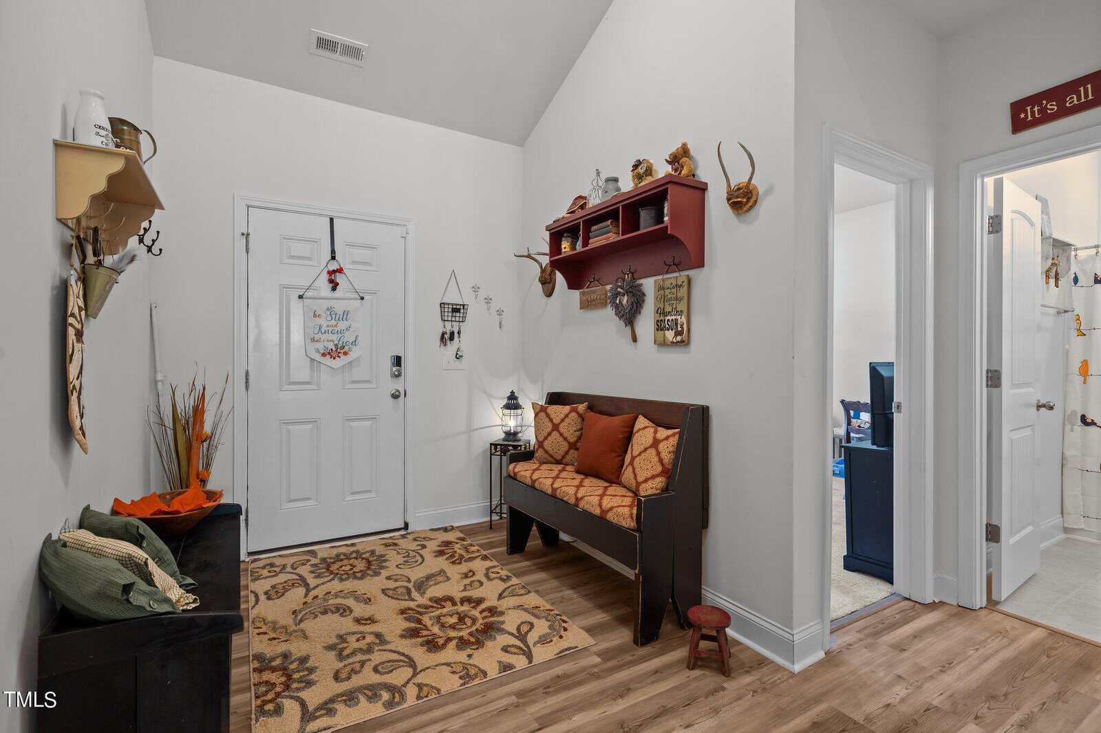 A small entryway with a white front door, a brown cushioned wooden bench with pillows, a decorative rug, wall shelves, and various wall-mounted decor items including antlers and hooks. A hallway is visible to the right.