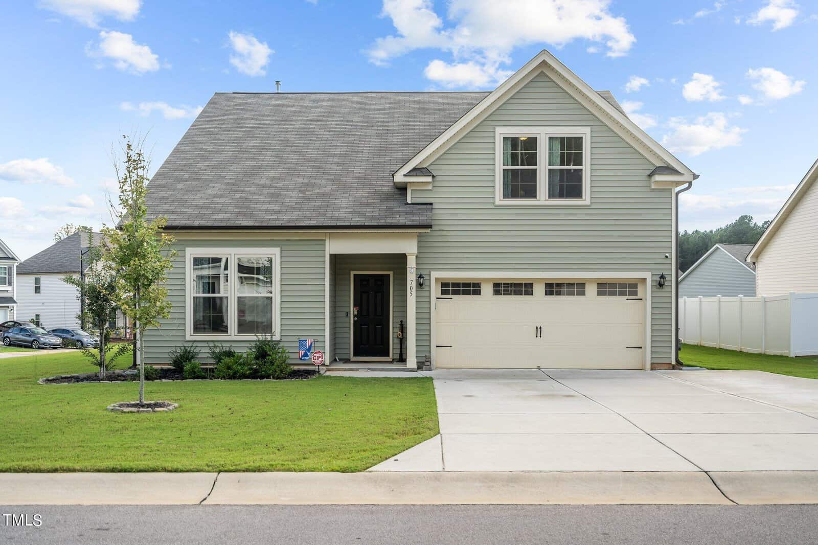 A two-story light gray house with white trim, a covered front porch, and an attached two-car garage. The lawn is neatly trimmed, and a small tree is planted beside a concrete driveway. The sky is partly cloudy.