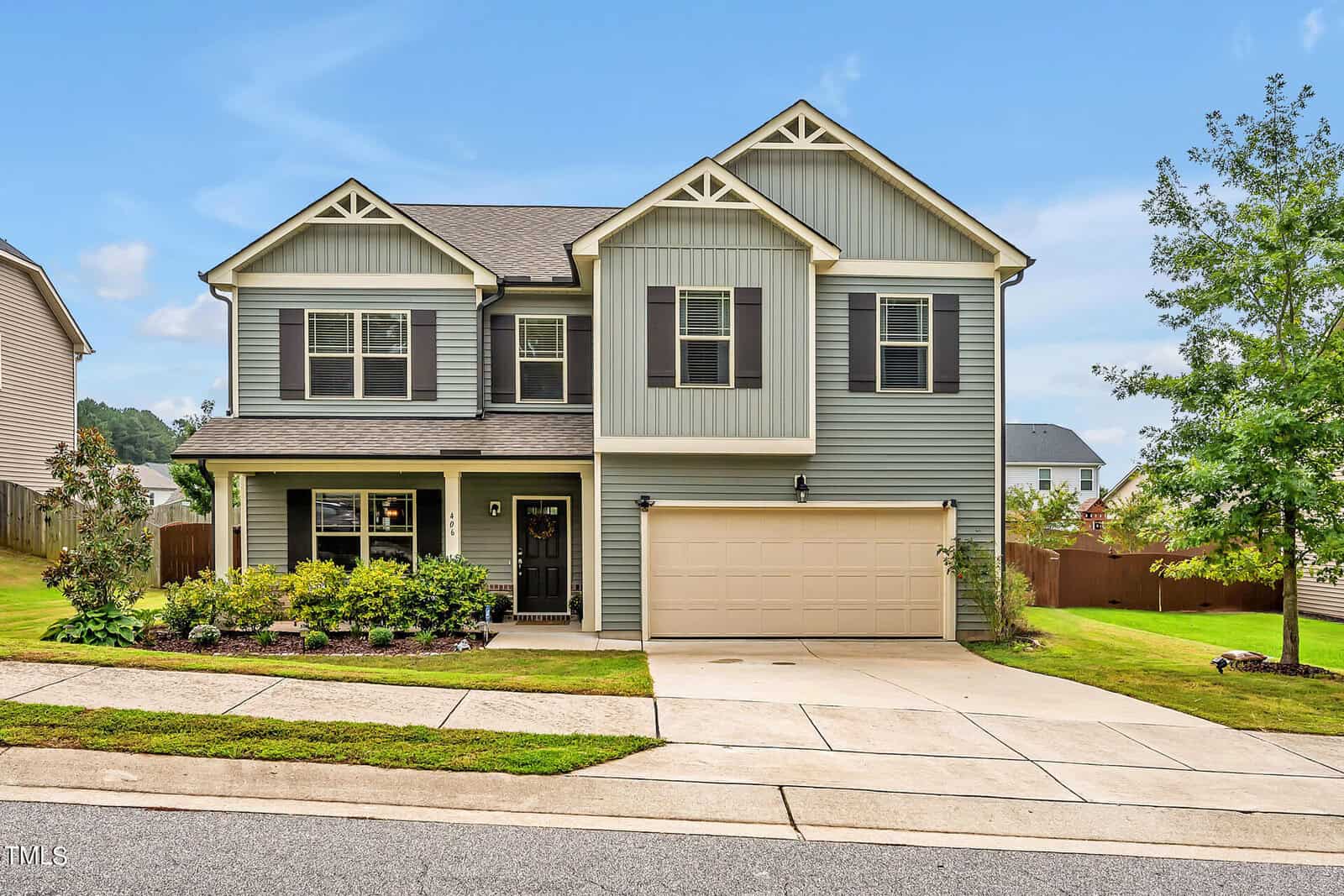 A two-story suburban house with light blue siding, dark shutters, a double garage, manicured lawn, and small shrubs in front. The house sits on a sloped driveway with a fenced backyard.