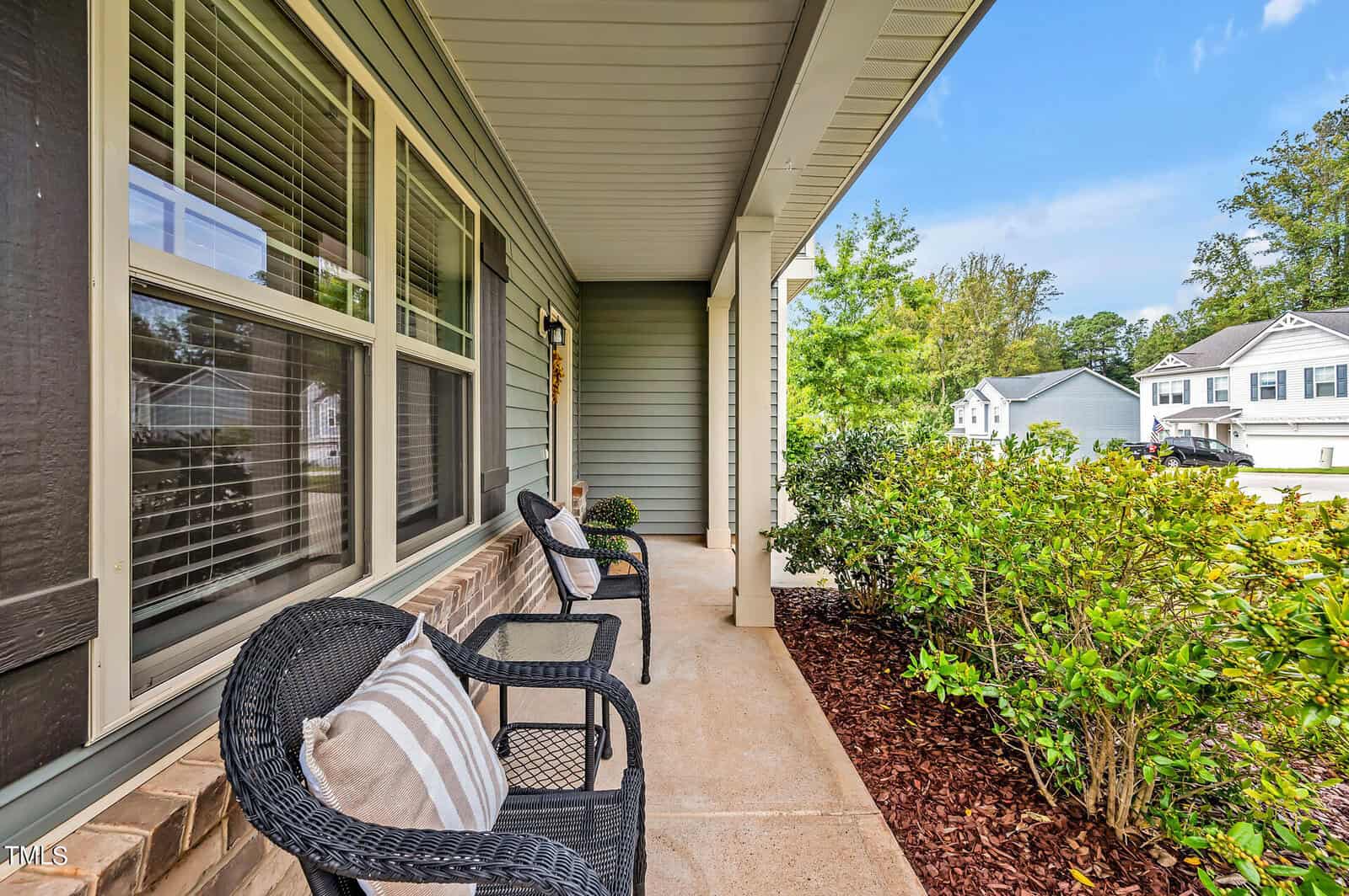 A covered front porch with two black wicker chairs, a small side table, tan brick trim, green shrubs along the walkway, and neighboring houses visible across the street under a blue sky.